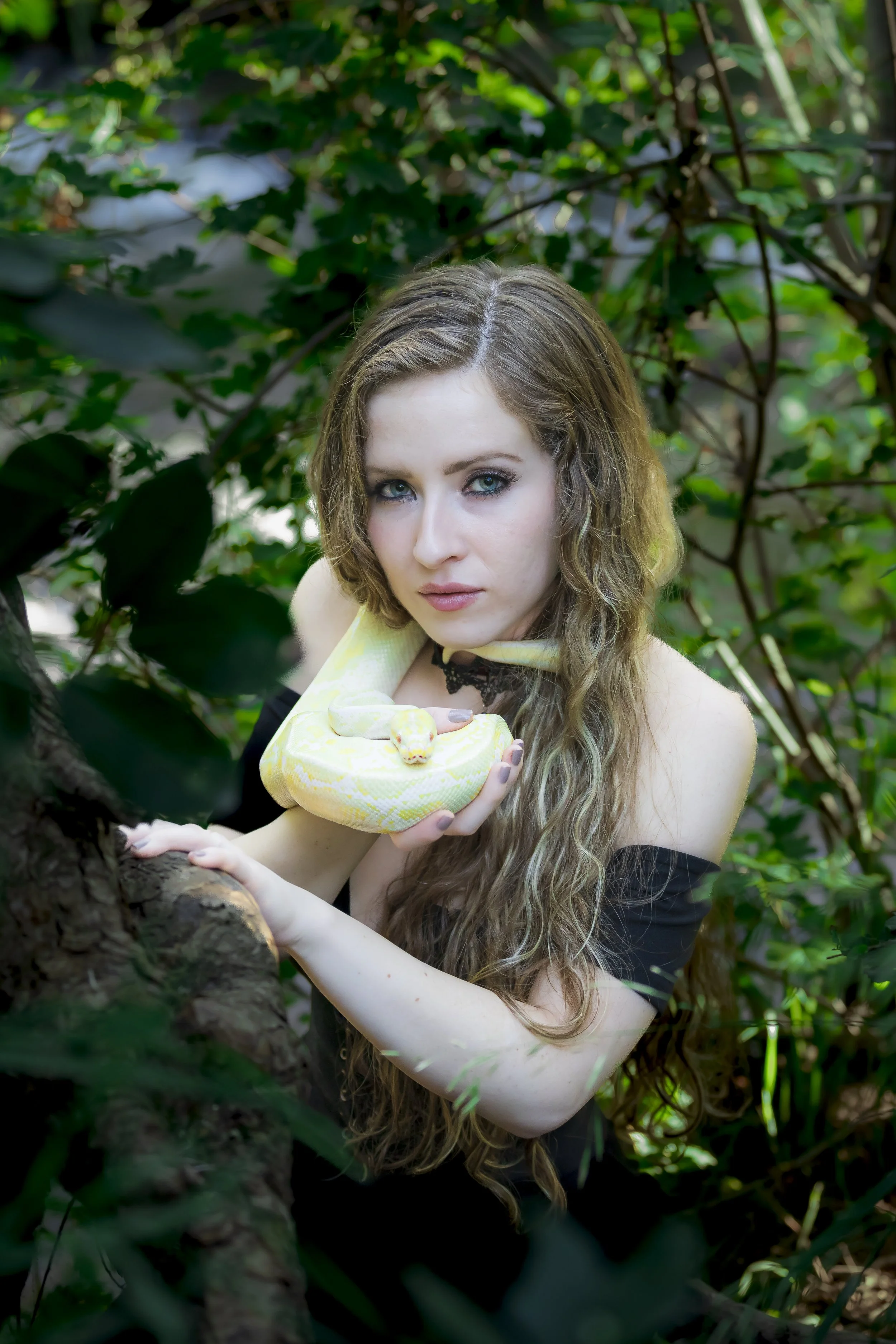 A woman with long, curly hair holds a yellow and white snake, with a green-yellow pattern, around her neck and on her hand, in a lush, green forest setting.