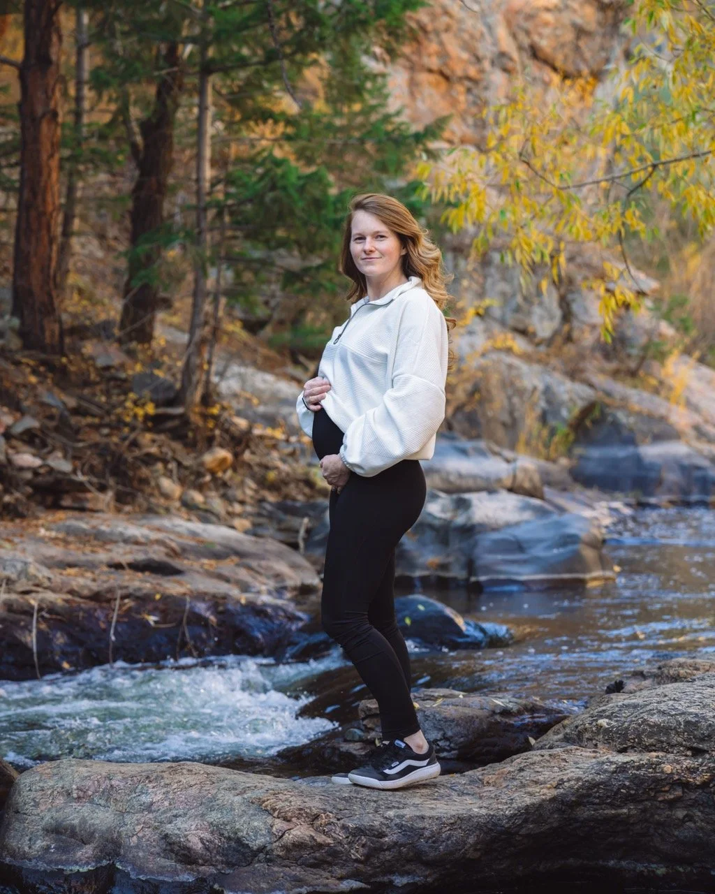 A woman standing on rocks in a shallow creek surrounded by trees with autumn foliage.