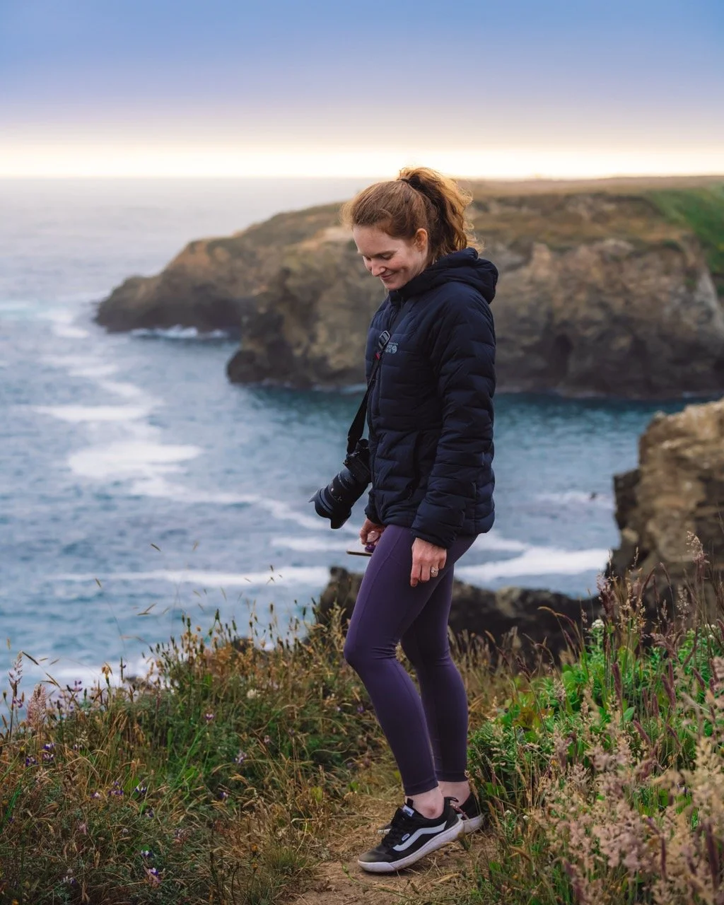 A woman in athletic clothing standing on a coastal trail with cliffs and ocean in the background during sunset, holding a camera and smiling.