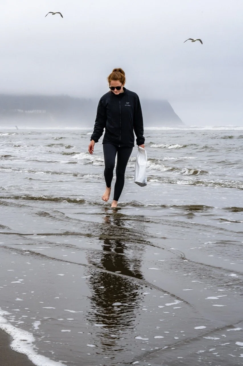 A woman in black jacket and leggings walking barefoot on the beach in Oceanside, OR with overcast sky, seagulls flying overhead, and water reflecting her image.