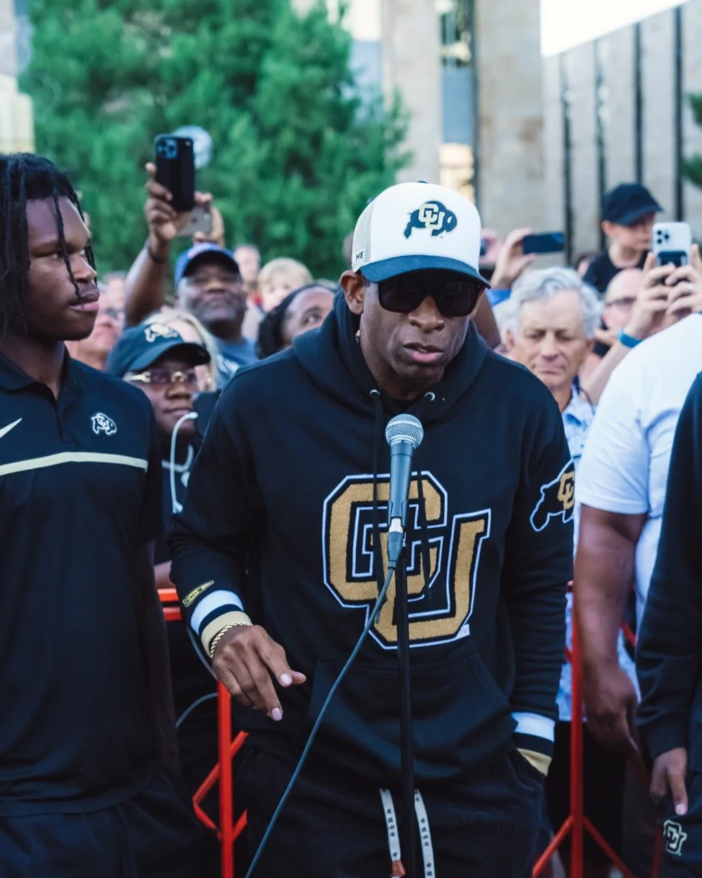 University of Colorado Football Head Coach Deion Sanders wearing a black hoodie and a white trucker cap with a logo, sunglasses, and a gold watch, standing in front of the Friday Night crowd in Boulder and speaking into a microphone.