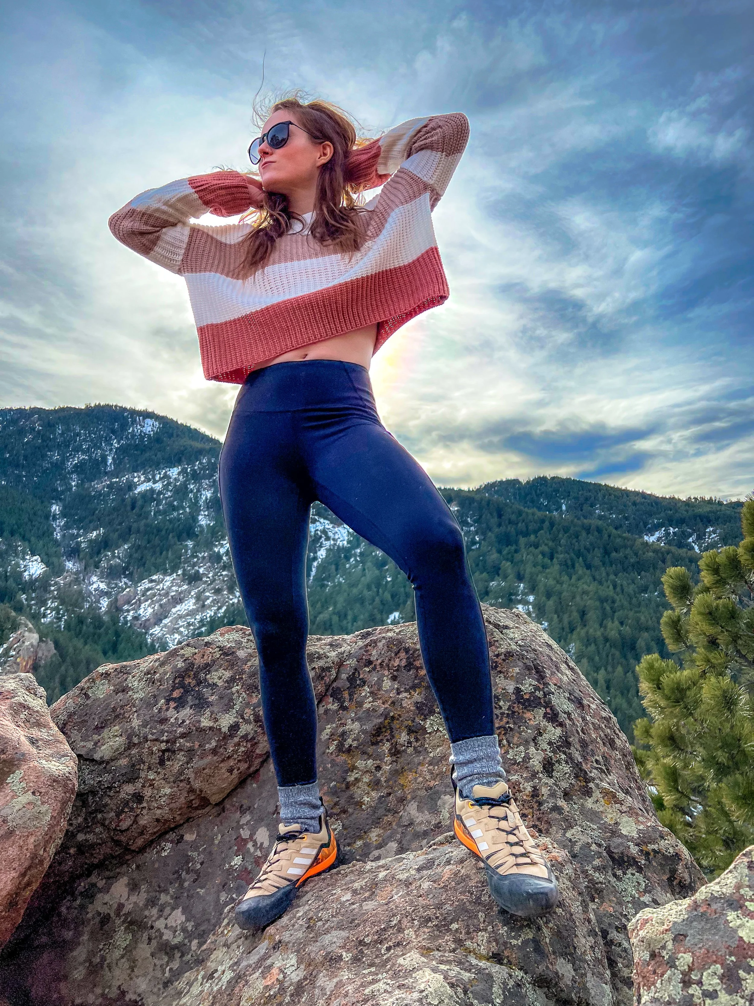 Woman standing on large rocks outdoors with mountain scenery, wearing sunglasses, a striped sweater, black leggings, and hiking shoes, with her hands behind her head, looking off into the distance.