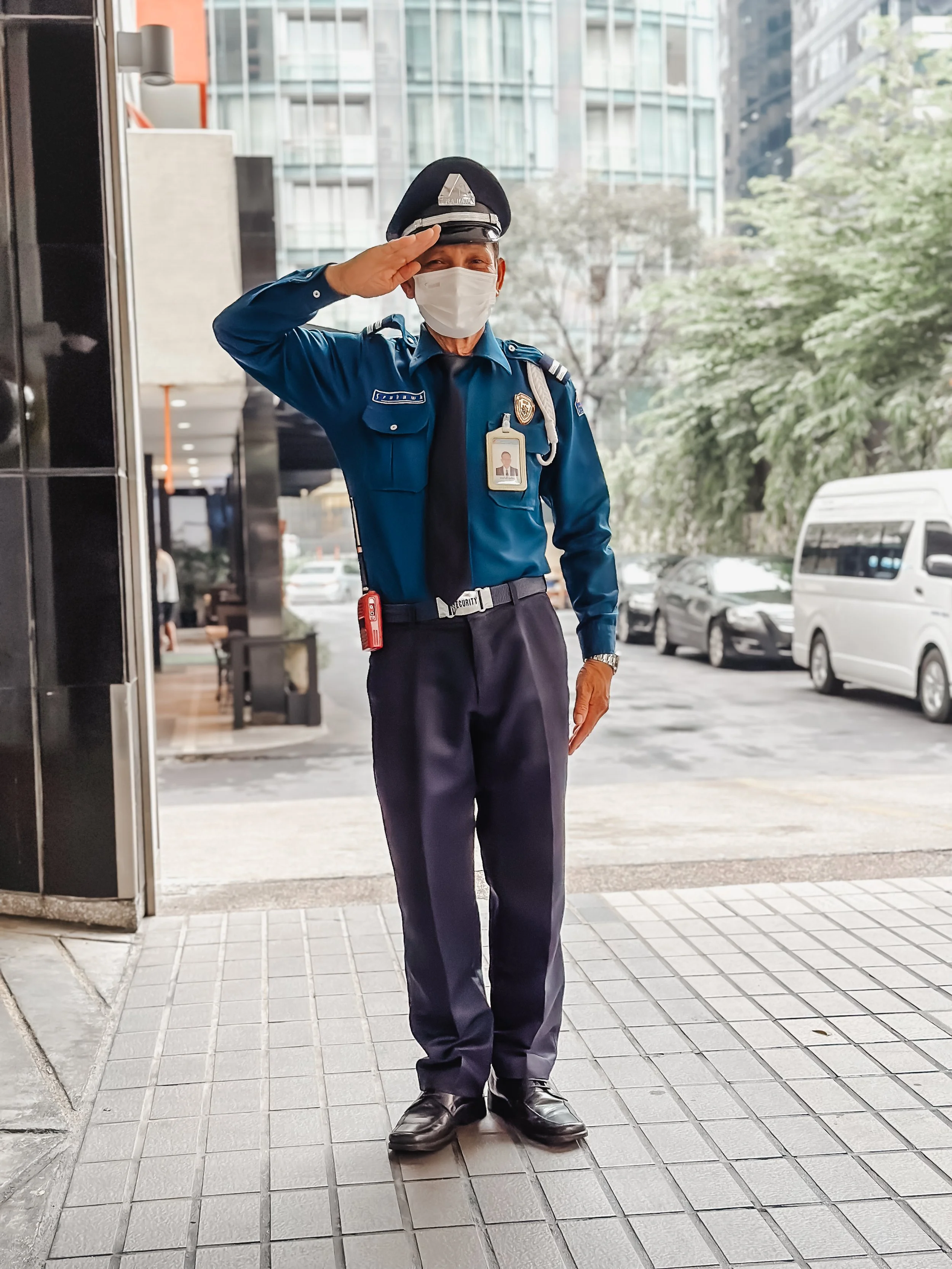 A Bangkok, Thailand security officer in uniform saluting outside a building with city street and parked cars in the background.