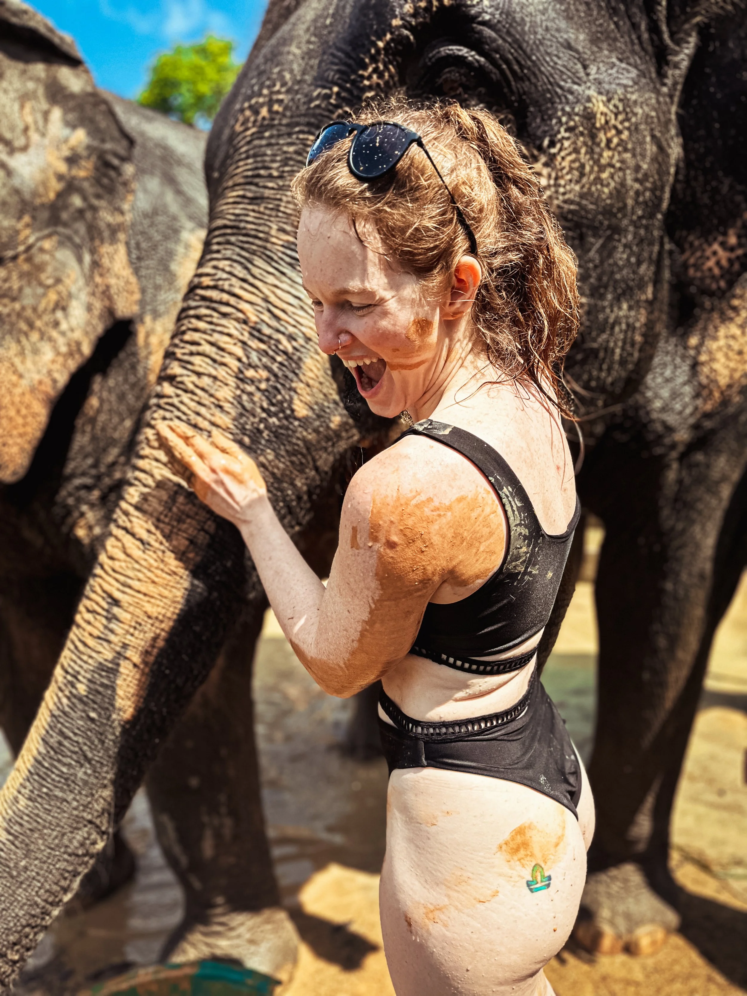 A woman in black clothing and sunglasses on her head is smiling and muddy from playing with elephants in Thailand, with one elephant in the background.