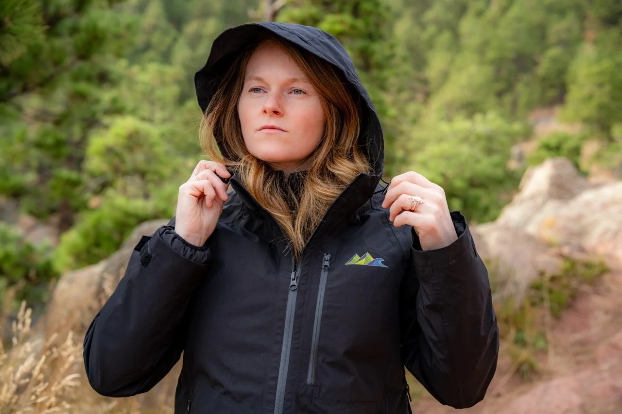 Woman in black outdoor jacket with hood, standing outdoors in a wooded area.
