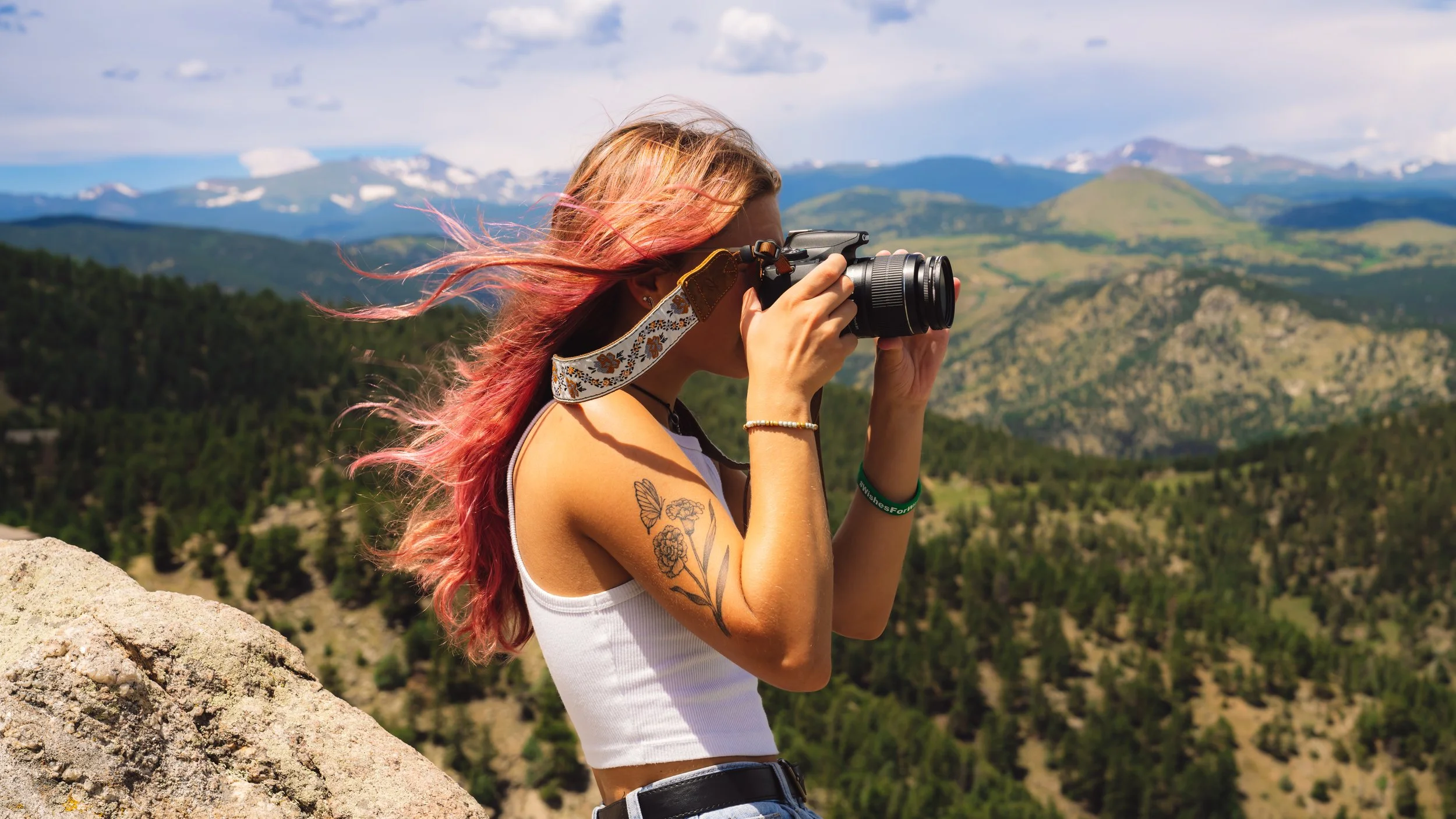 Woman with pink hair taking a photograph with a camera on a mountain overlook with green hills and snow-capped mountains in the background.