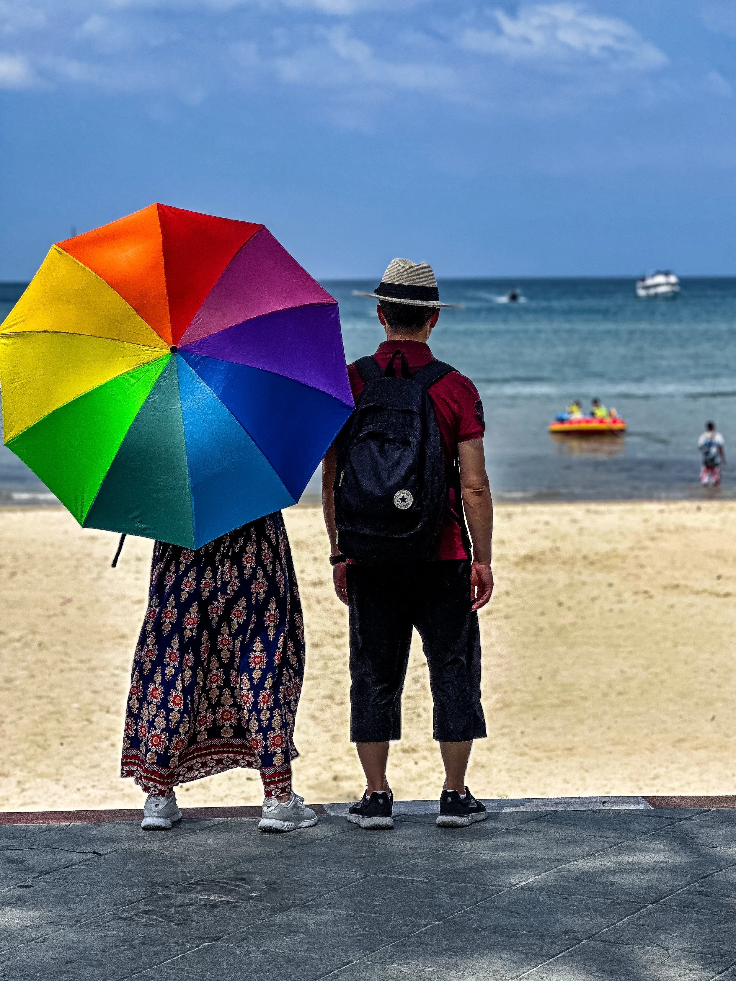 A couple stands on a beach in Phuket, Thailand, with the woman holding a rainbow umbrella, facing the ocean. The man wears a fedora, red polo shirt, and backpack, and they watch boats pass by on the water.
