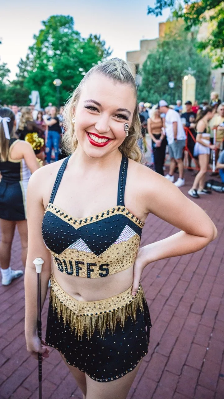 A University of Colorado Featured Twirler with blonde hair smiling at a parade in Boulder, wearing a cheerleading uniform with the word 'BUFFS' on it, black and gold colors, and holding a white baton.