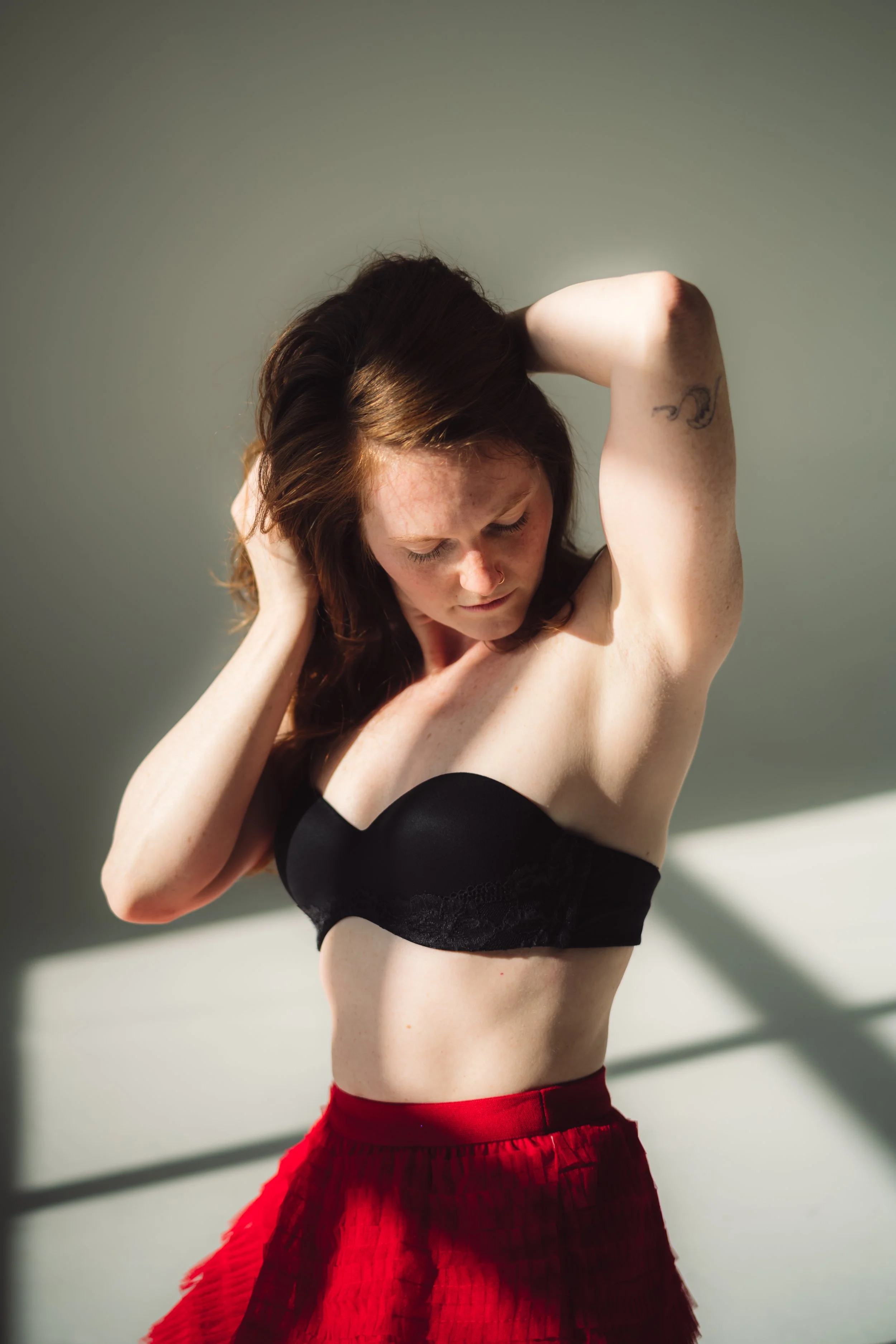 A woman with red hair, wearing a black strapless top and red skirt, standing in sunlight with her eyes closed, touching her neck and head.
