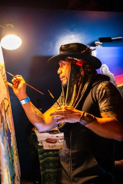 A male artist wearing a hat and scarf painting on a canvas in a dimly lit room.