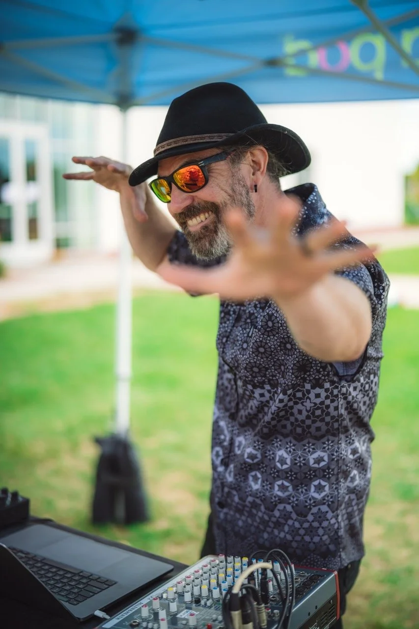 Random Rab smiling with a beard, wearing a black hat, sunglasses with orange reflective lenses, and a patterned shirt, reaching towards the camera with his arms extended.