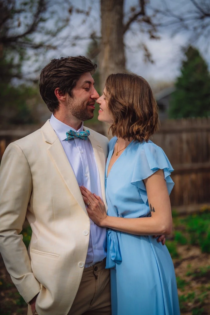 A couple stands close in a backyard, about to kiss, with a large tree and wooden fence in the background.
