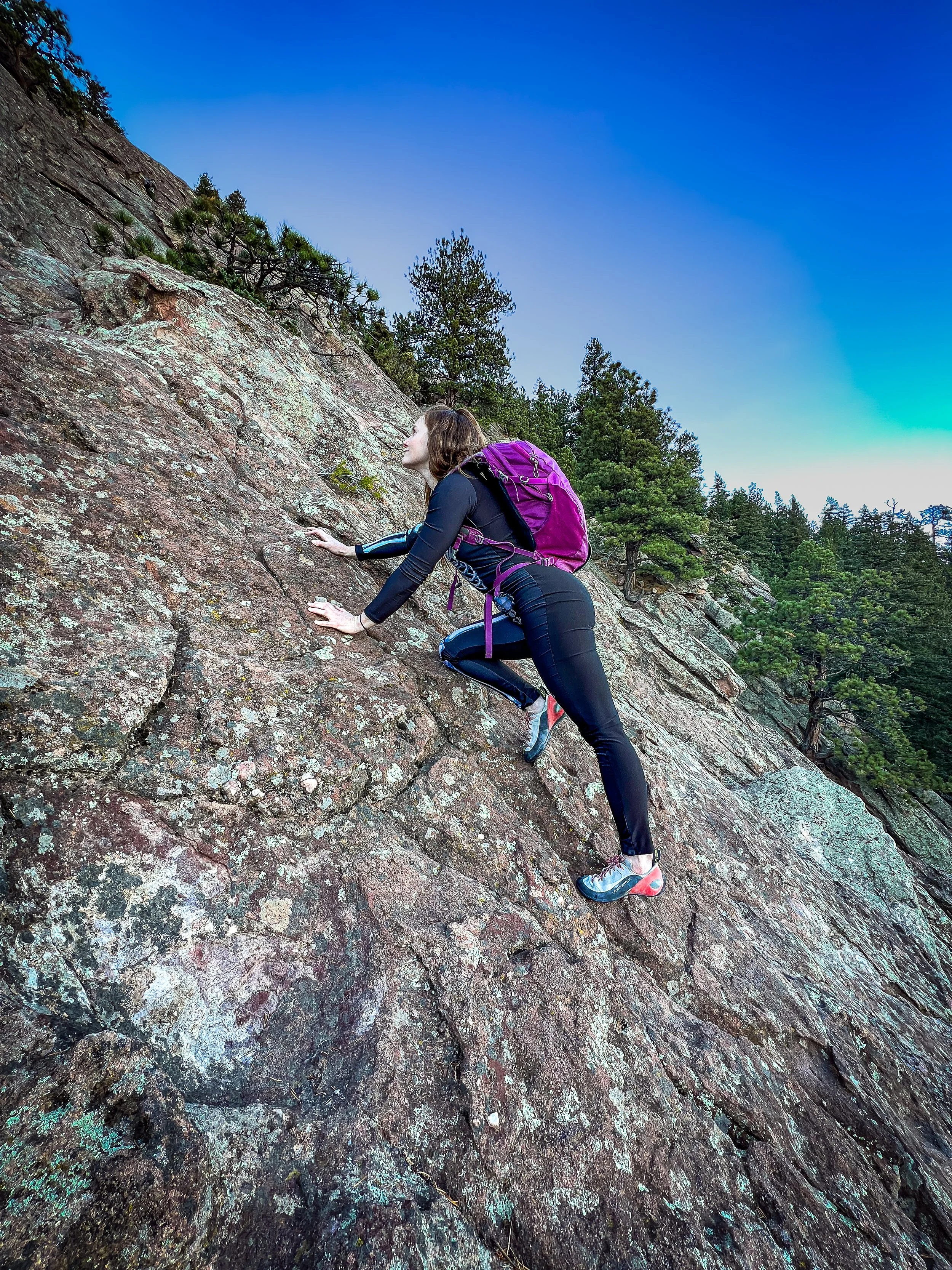 Woman in black athletic clothes climbing a rock face outdoors with a purple backpack and mountain landscape in the background.