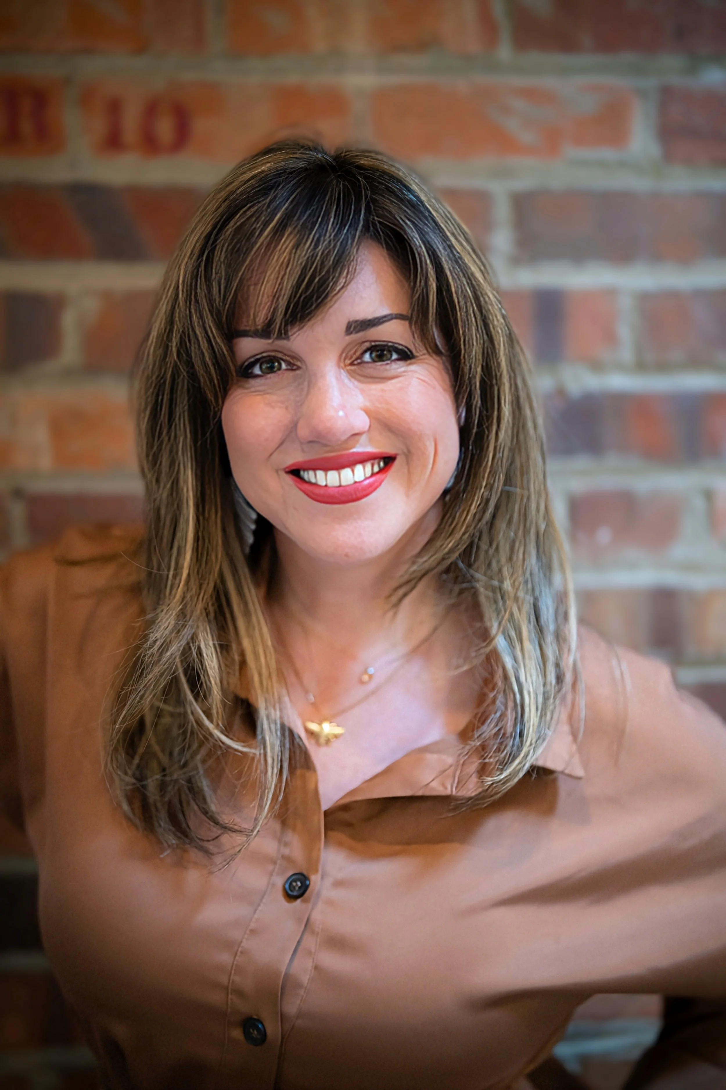 A woman with shoulder-length brown hair and light skin smiling at the camera, wearing a brown shirt and a gold butterfly necklace, with a brick wall in the background.