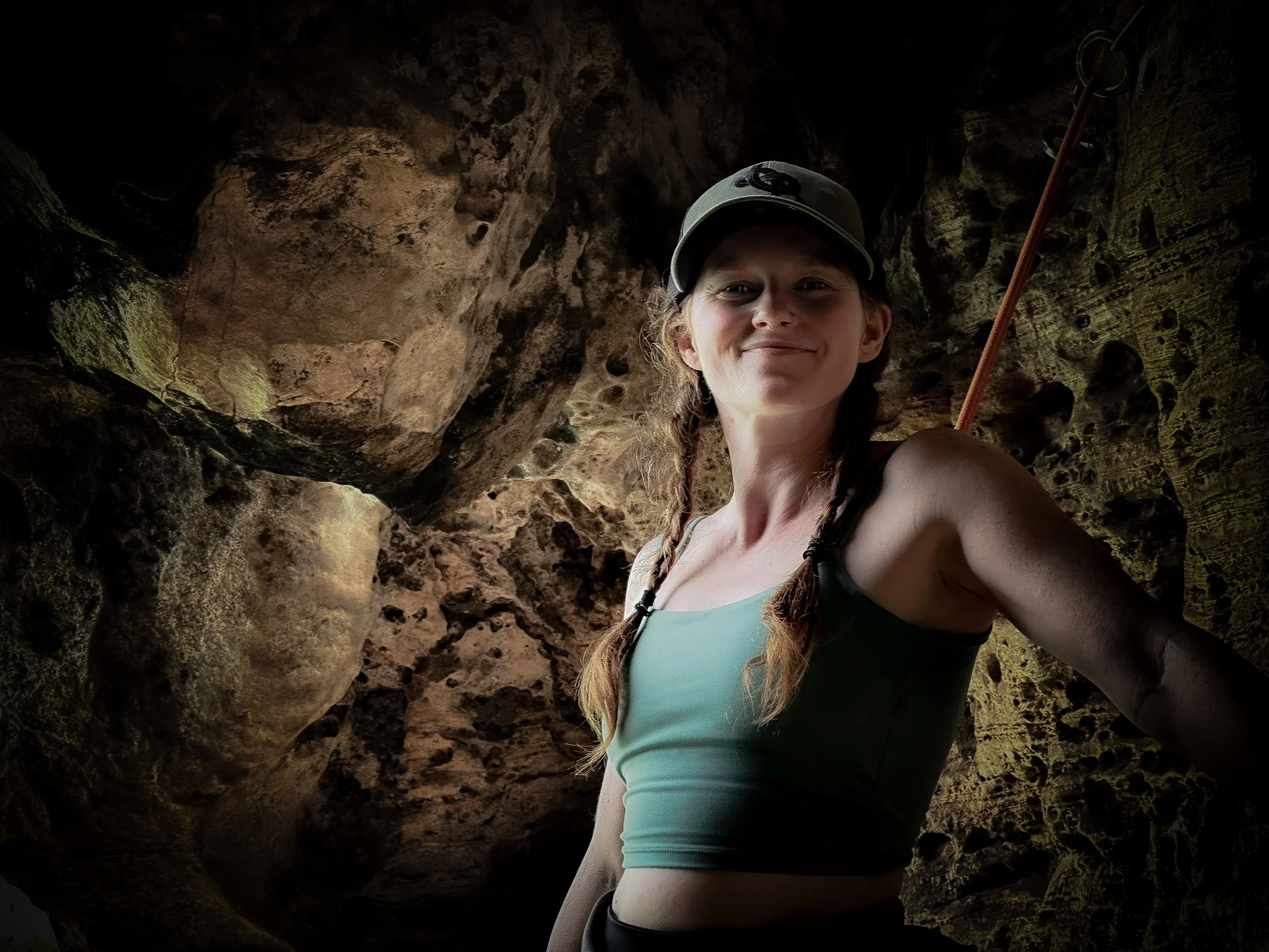 A young woman with braids, wearing a gray cap and a green sports top, stands inside a cave with rocky walls, smiling at the camera.