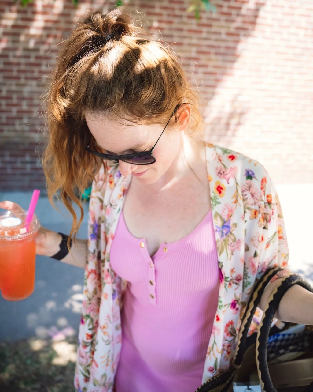 A young woman with wavy red hair and wearing sunglasses is outdoors, wearing a pink top and a floral jacket. She is holding a pink beverage with a pink straw in her right hand and appears to be looking down.