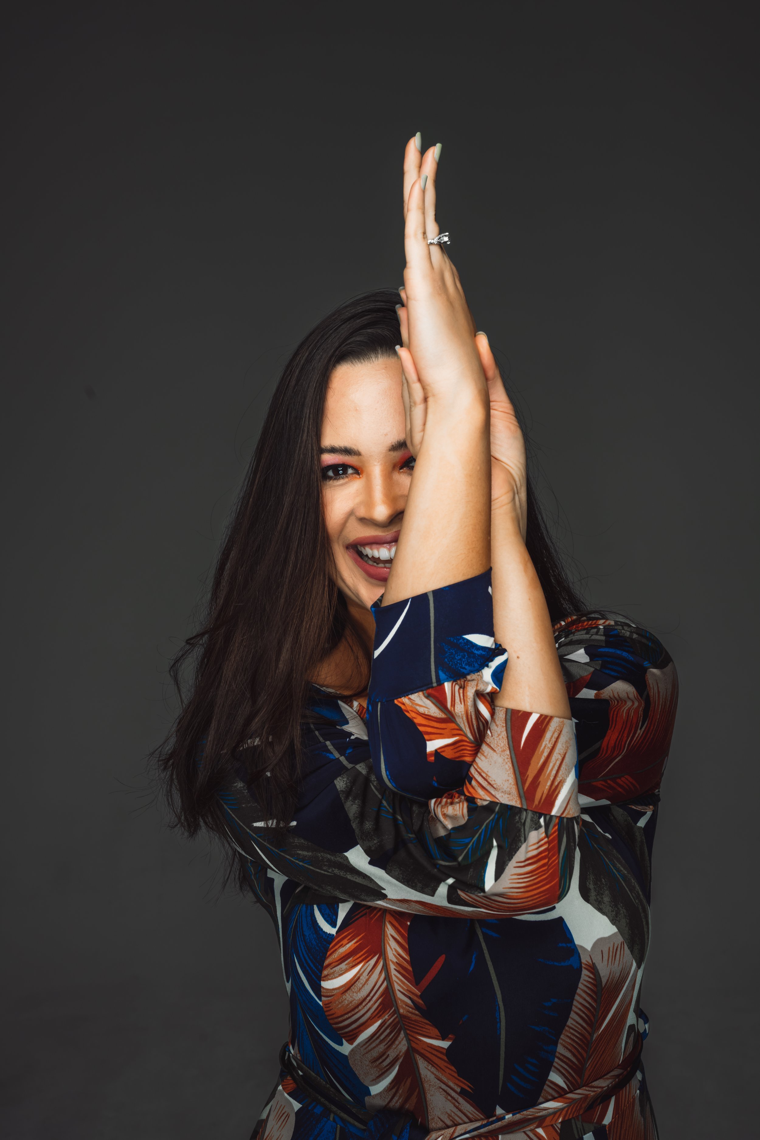 A woman with long dark hair smiling and holding her hands together near her face against a dark background.