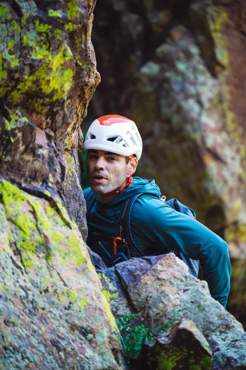 A man wearing a white helmet with red and green details, dressed in a blue jacket and backpack, bouldering on a moss-covered rock face outdoors surrounded by natural rock formations and greenery.