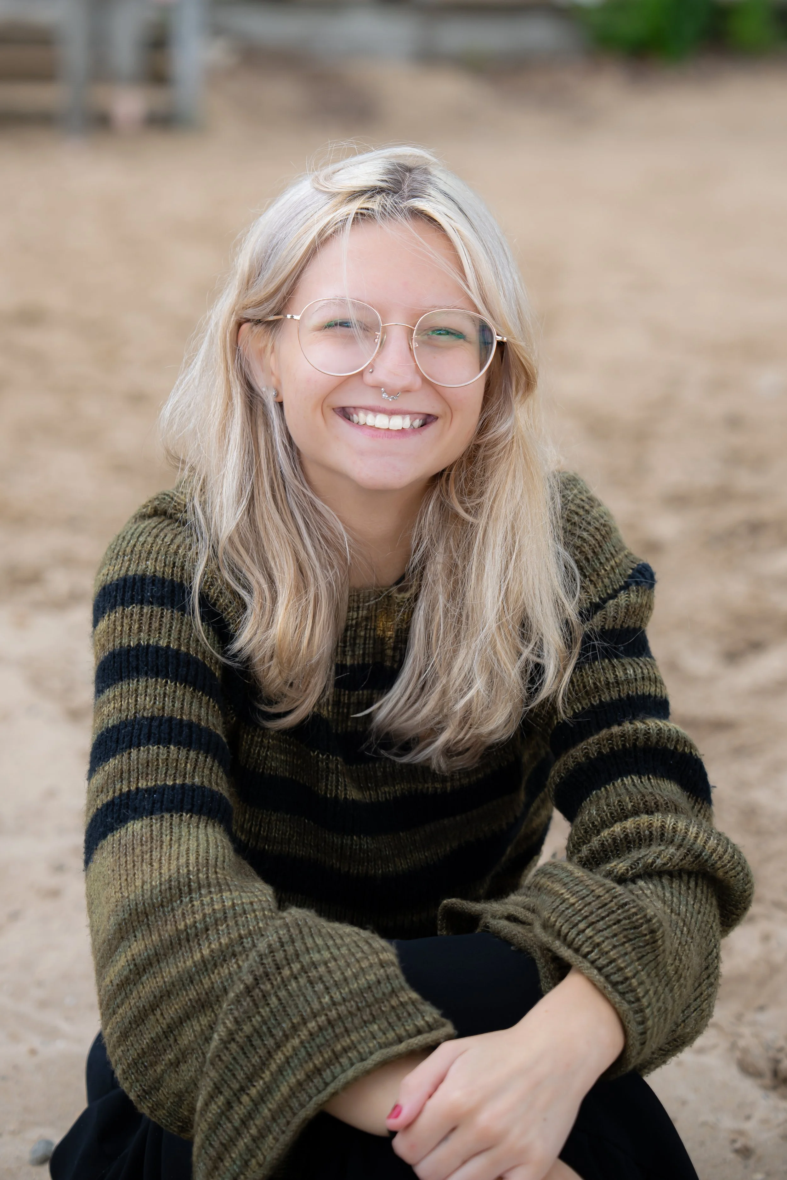A young woman with long blonde hair, glasses, and a septum piercing, smiling and sitting outdoors on a sandy surface, wearing a striped sweater.