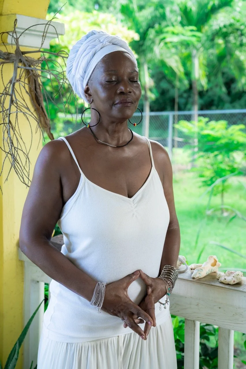 An older woman with dark skin and closed eyes, wearing a white headwrap, white tank top, and jewelry, standing in a lush green outdoor setting with a yellow pillar and decorative shells on a table nearby.