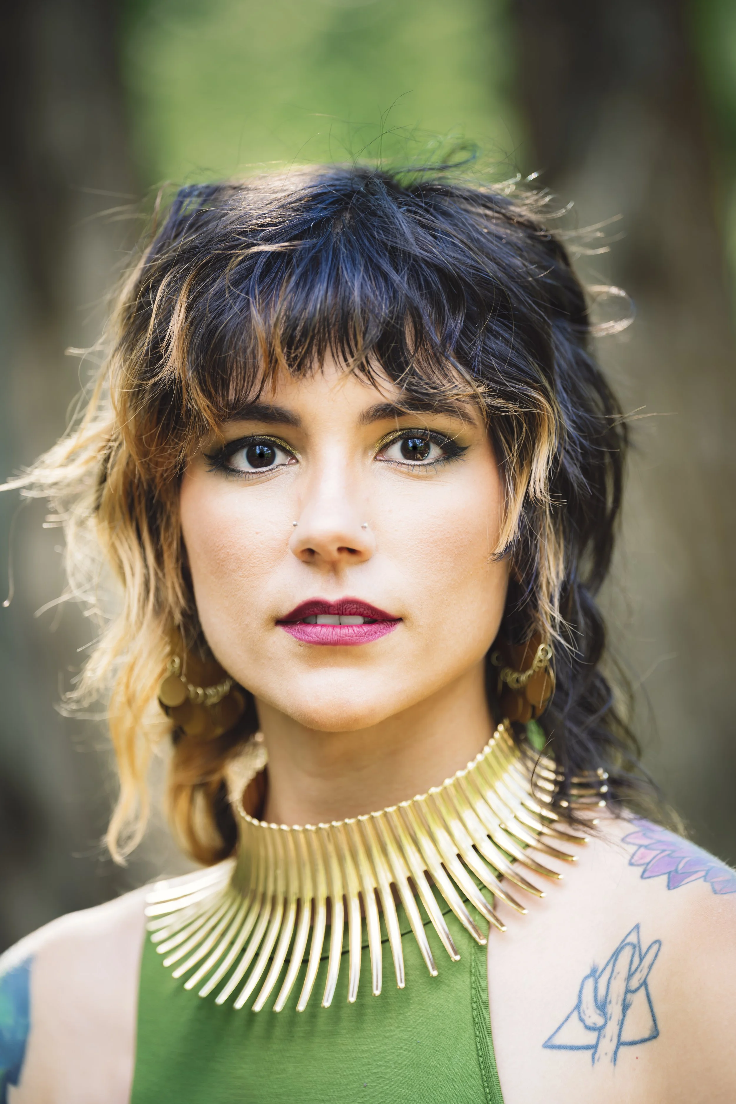 A woman with short, wavy black and blonde hair, wearing makeup with bold lipstick, and dressed in a green top with a large gold statement necklace and earrings. 