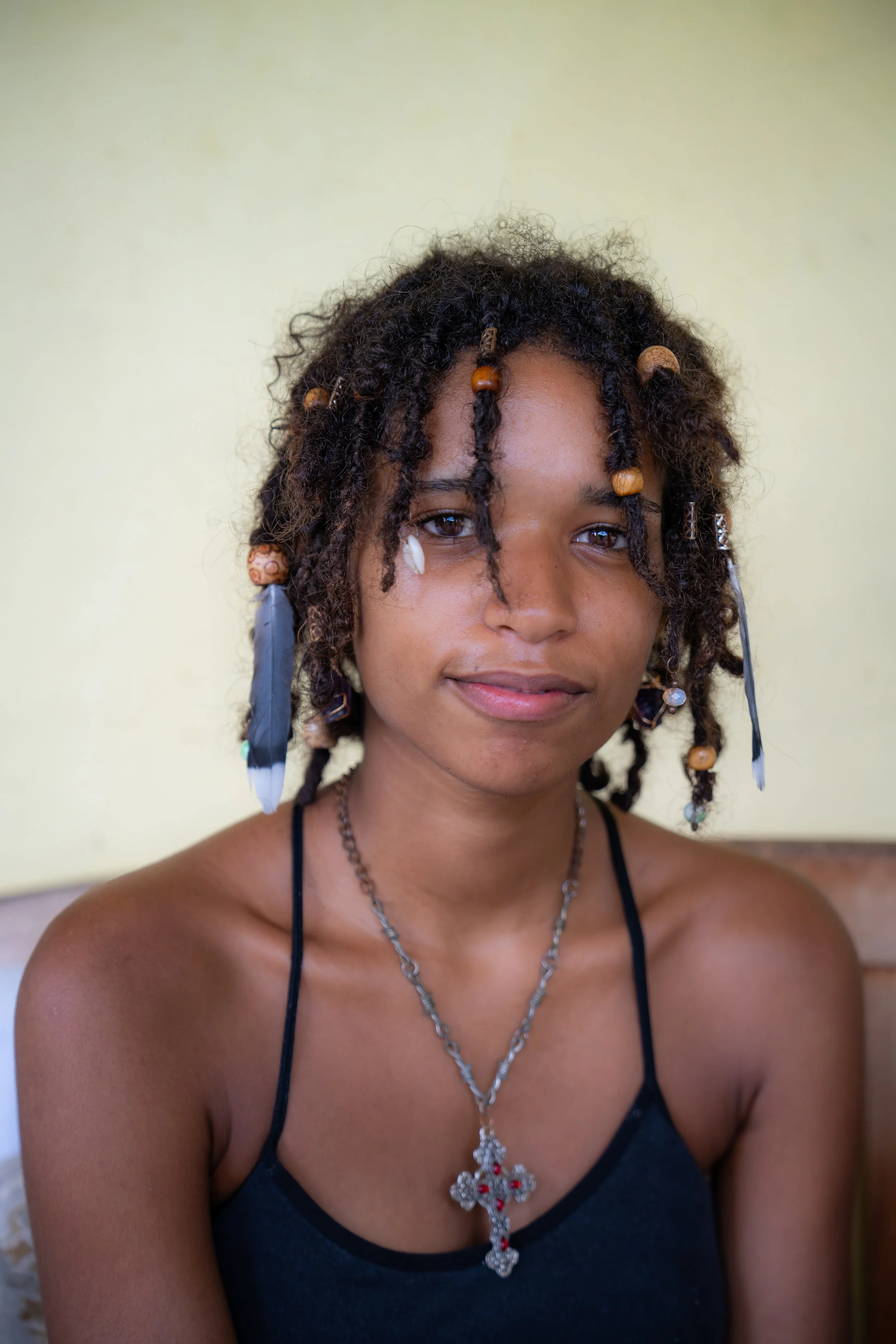 A young woman with curly hair decorated with beads and feathers, wearing a black tank top and a cross necklace, sitting indoors against a light yellow background.