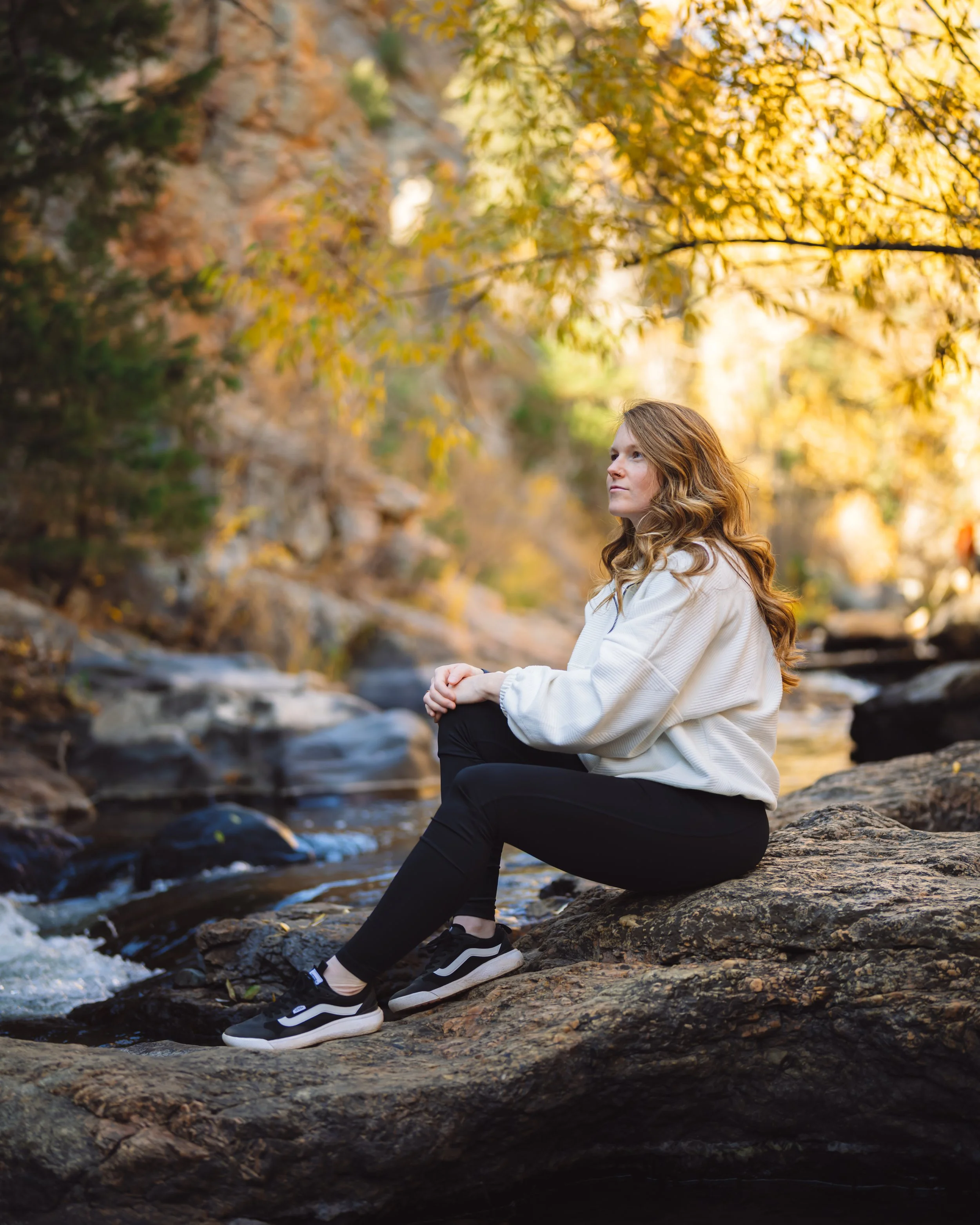 A woman with long red hair sits on a rock by a creek in Lair O' the Bear Park, CO surrounded by autumn trees with yellow leaves.