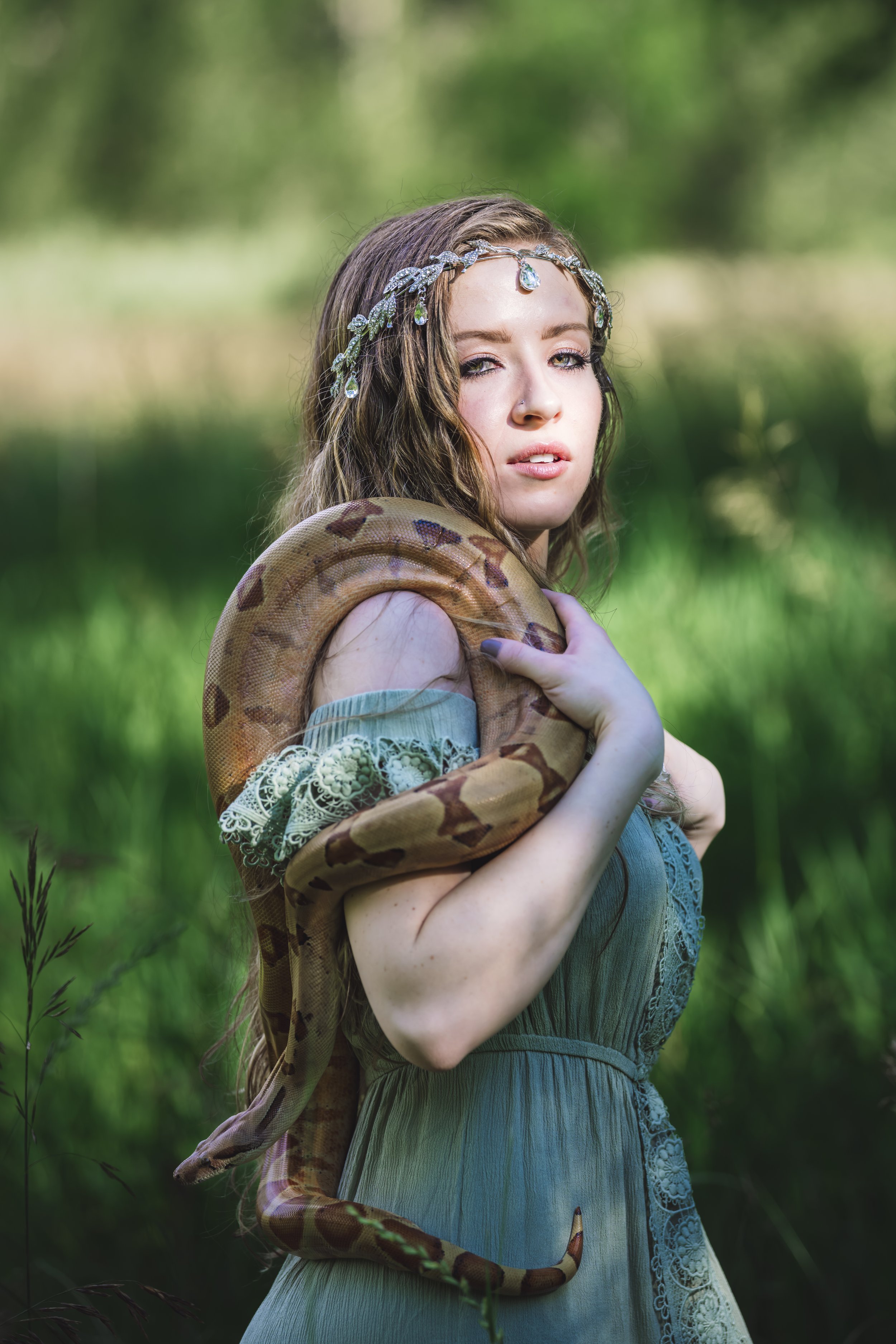 A young woman with long wavy brown hair, wearing a blue dress with lace details and a jeweled headband, is outdoors in a green forest, holding a large snake around her shoulders and neck.