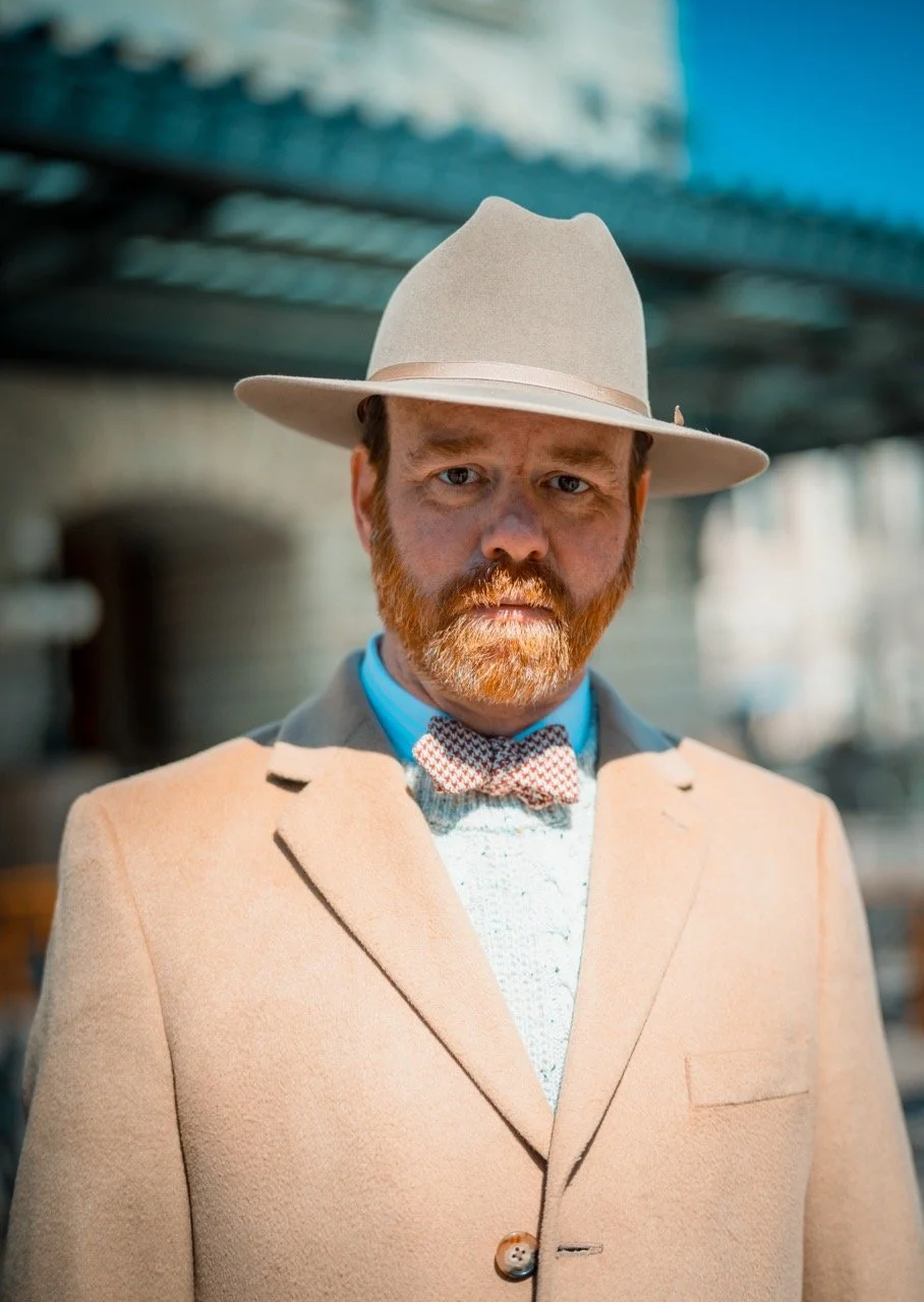 A man with a red beard wearing a beige coat, a white hat, a bow tie, and a sweater, standing outside with a blurred Denver Union Station in the background. 