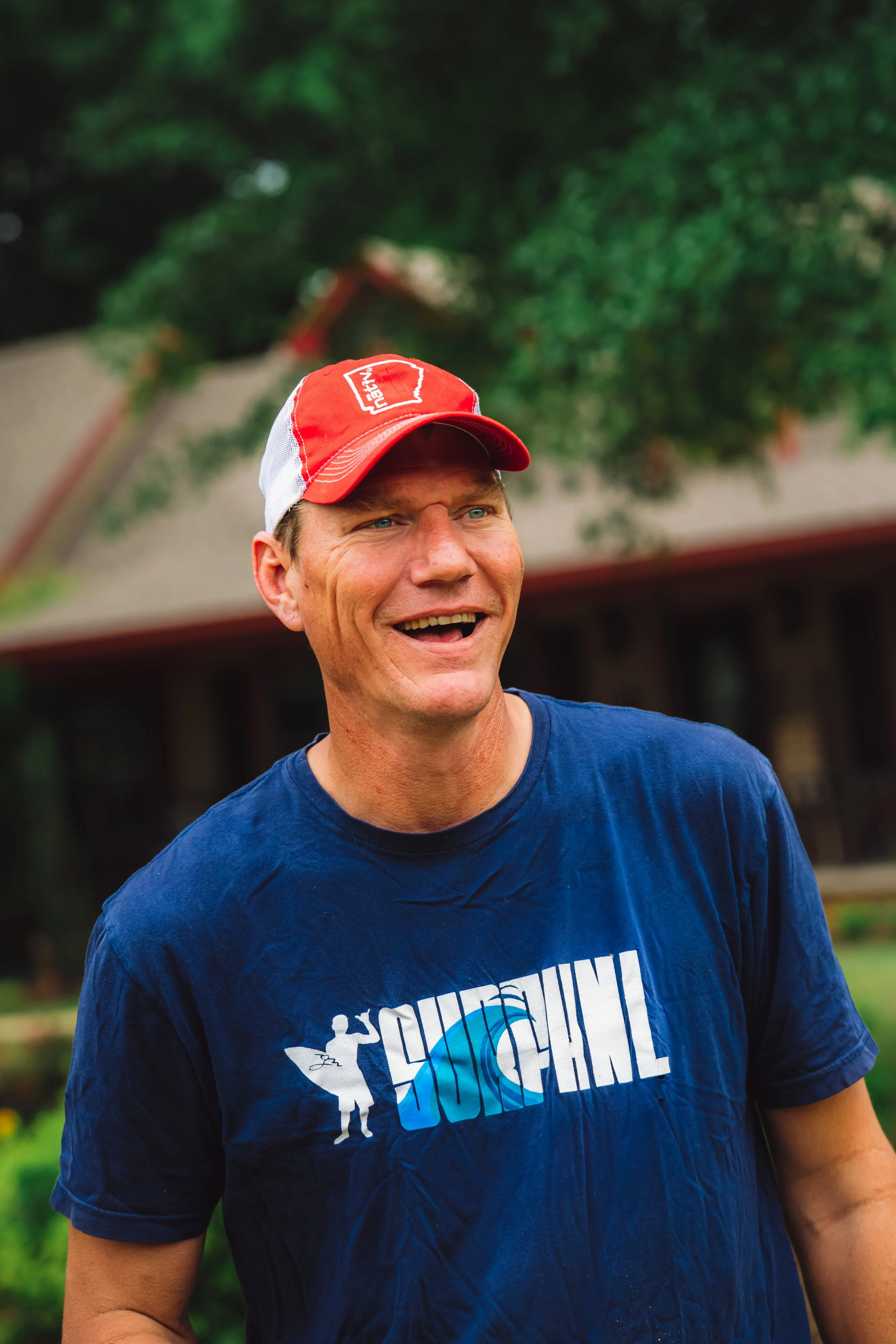 A man smiling outdoors, wearing a red and white cap and a blue T-shirt in front of a house and surrounded by green trees.