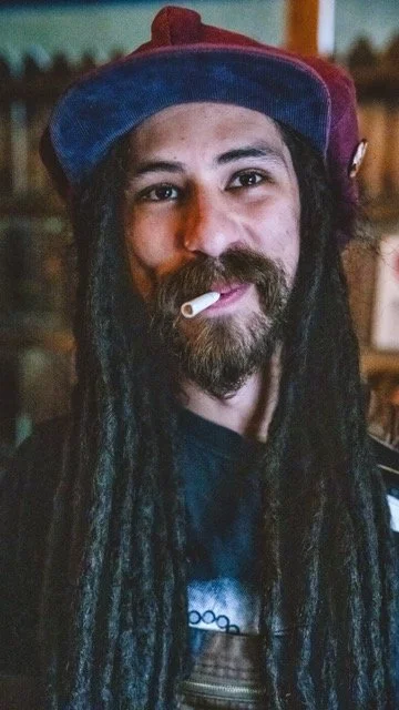 Man with long dreadlocks, a beard, wearing a multicolored cap, with an unlit cigarette in his mouth.