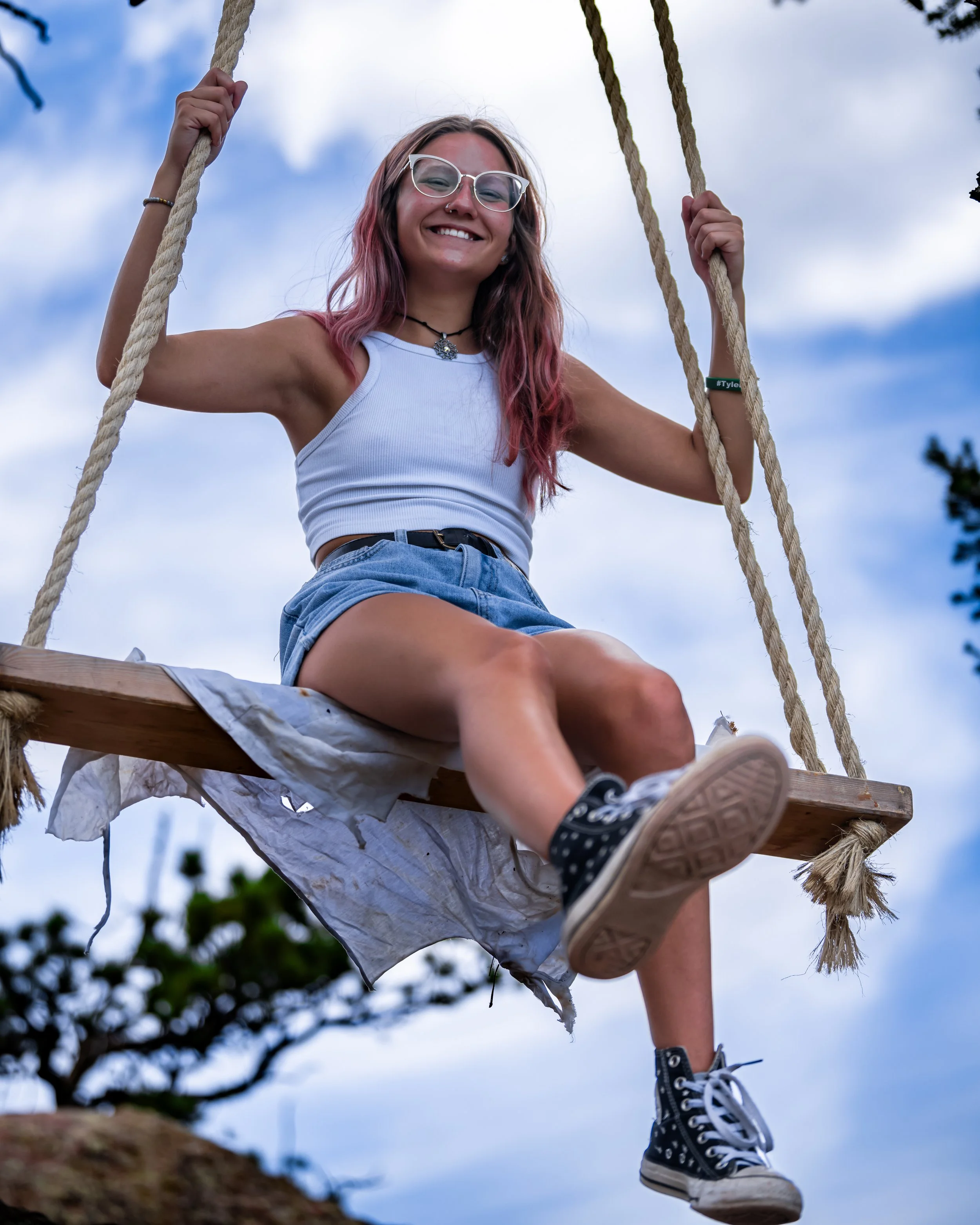 Young woman with glasses and pink hair smiling while sitting on a wooden swing outdoors, wearing a white crop top, denim shorts, and black polka dot sneakers, under a partly cloudy sky.