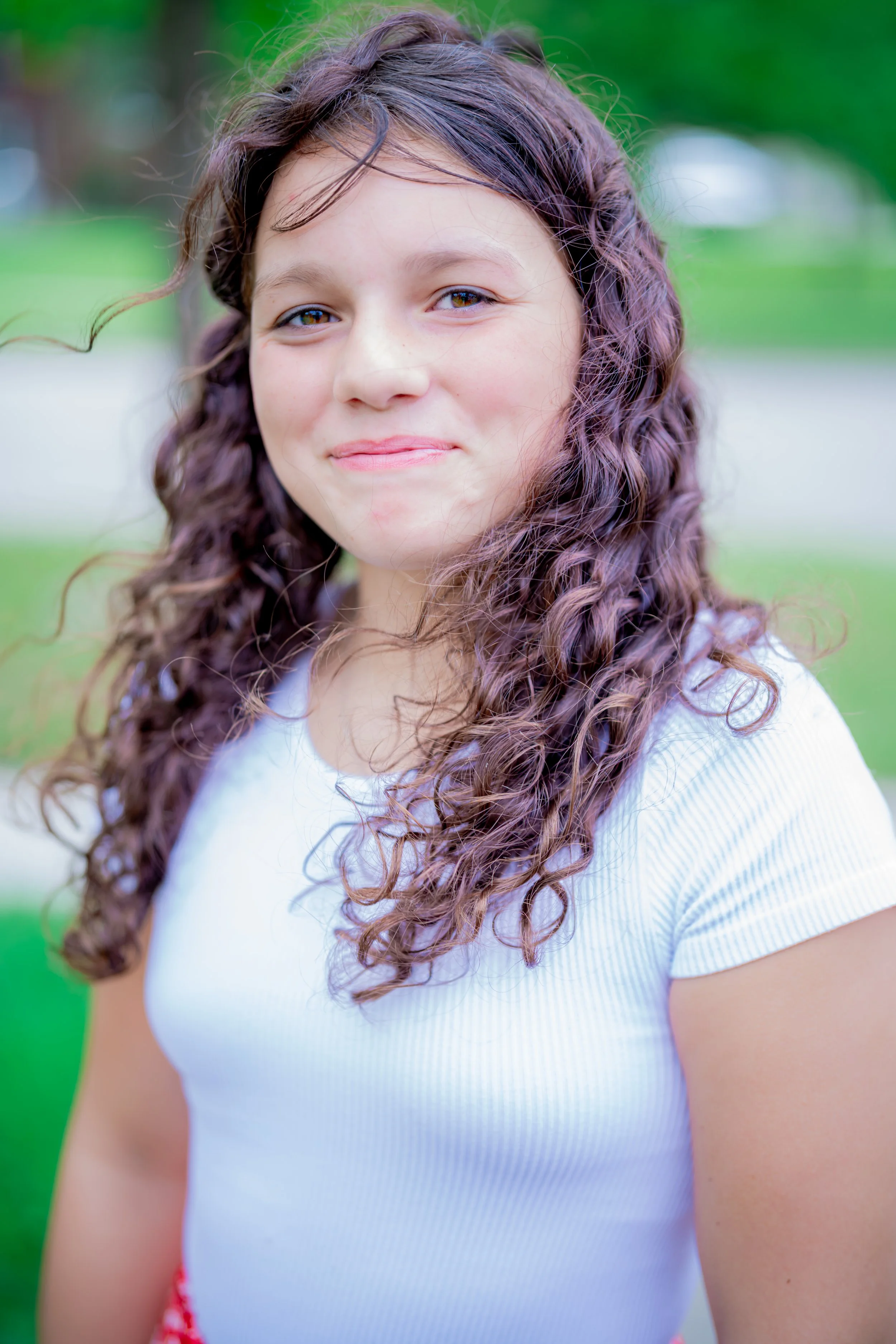 A young girl with long, curly hair smiling outdoors with a green background.