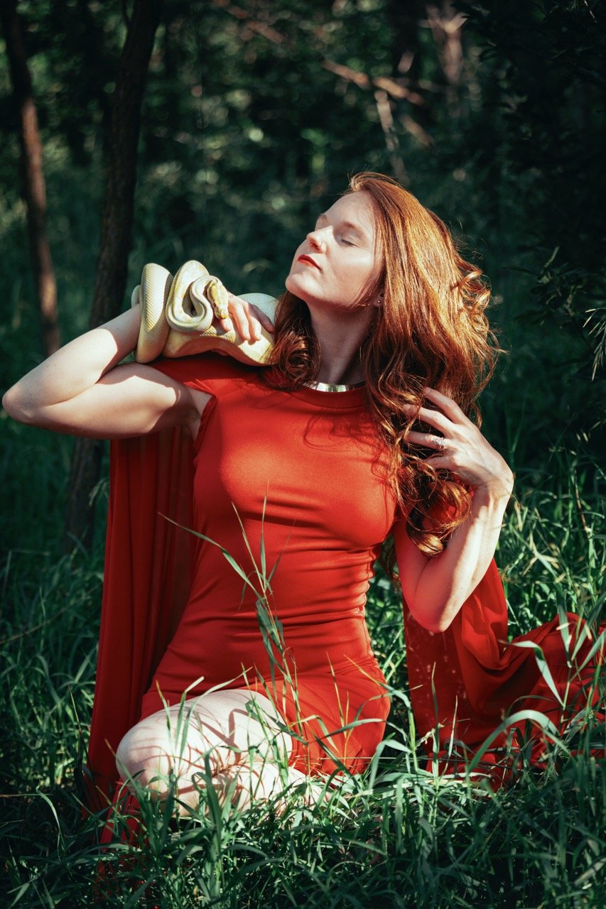A woman with red hair and a red dress kneeling in a grassy outdoor setting, holding a yellow snake on her shoulder with her eyes closed in a serene pose.