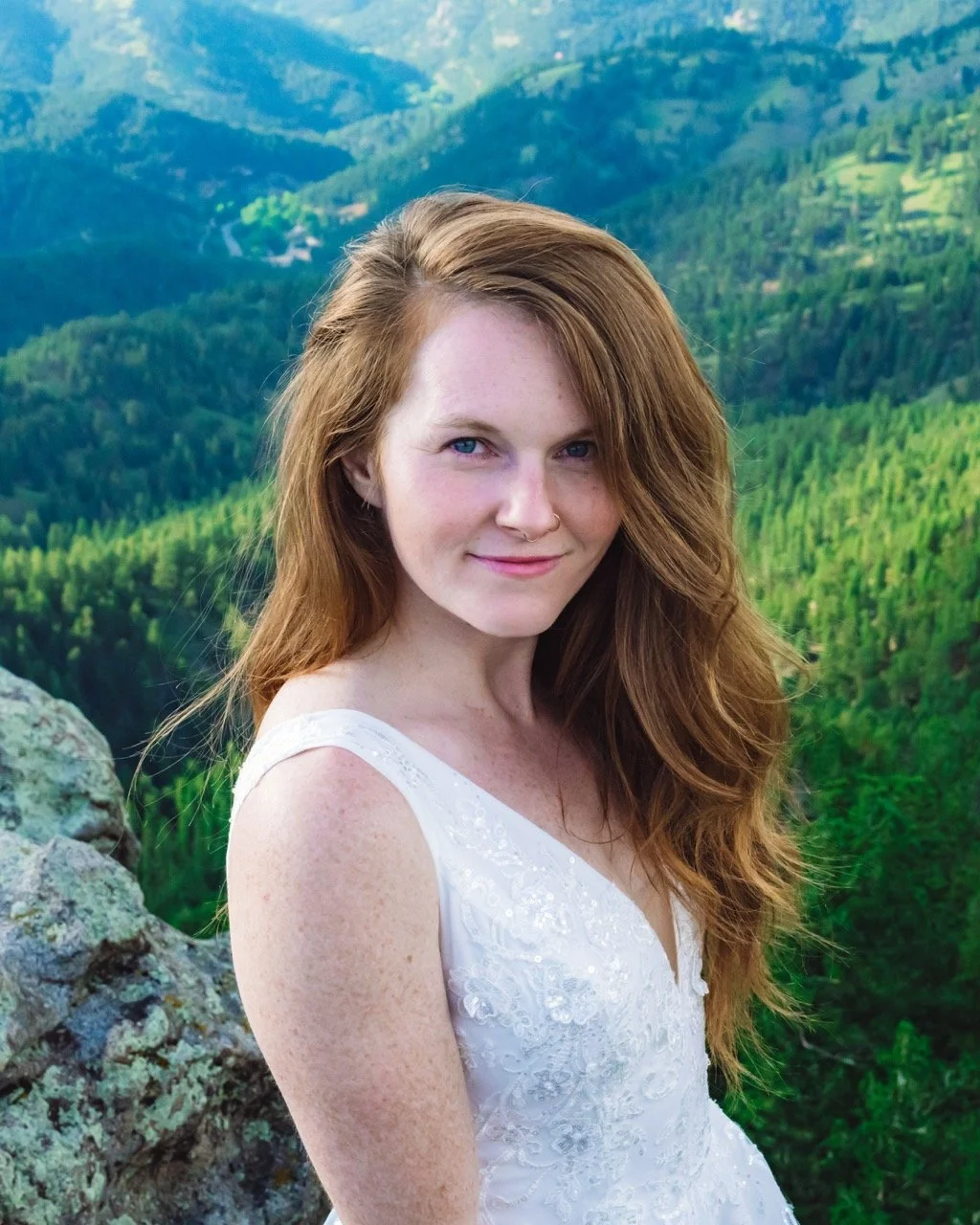 A woman with long red hair, wearing a white dress, standing outdoors with a mountain landscape in the background.