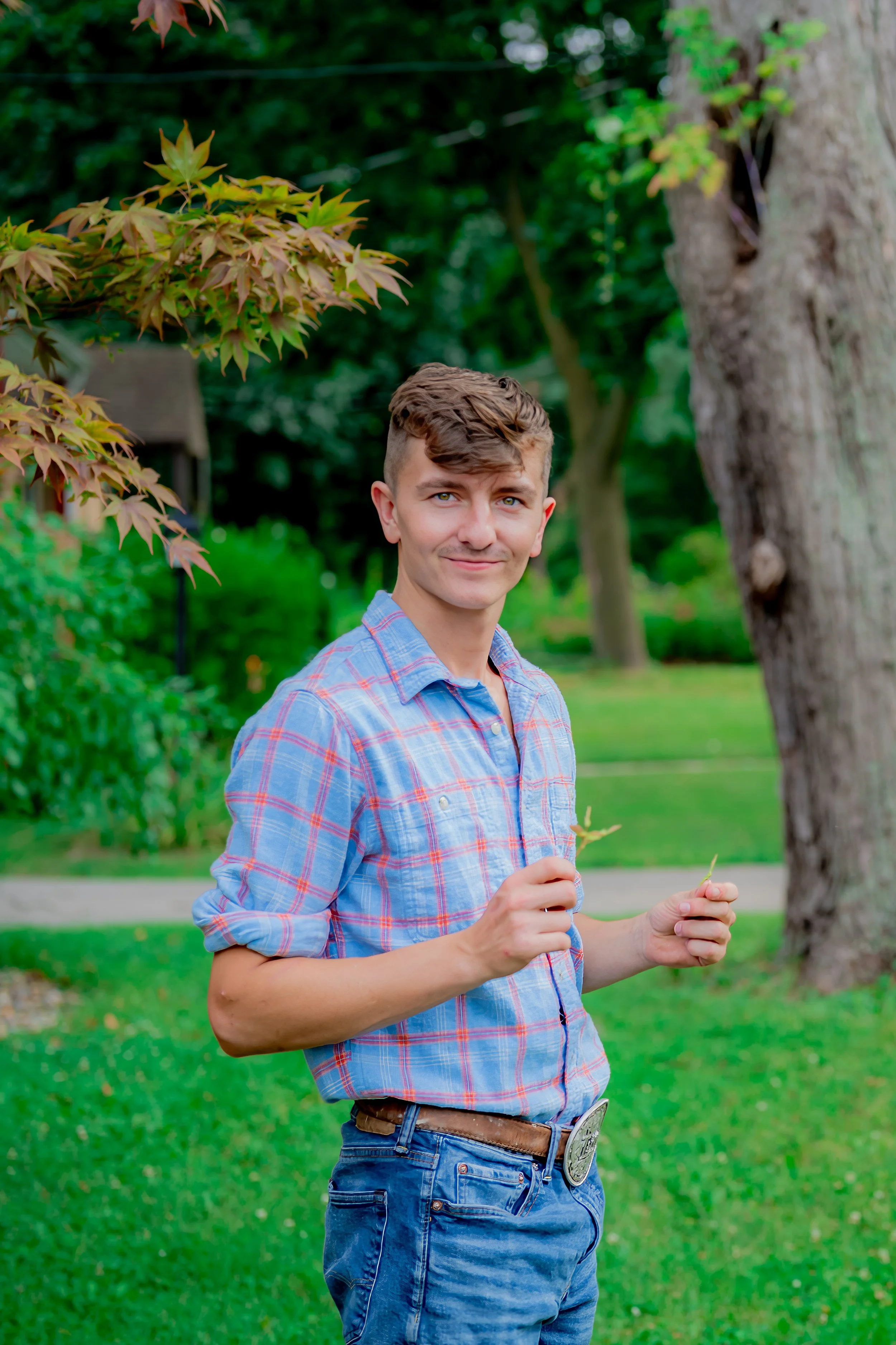 A young man in a plaid blue shirt and jeans stands outdoors in a park, holding small branches, with green trees and grass in the background.