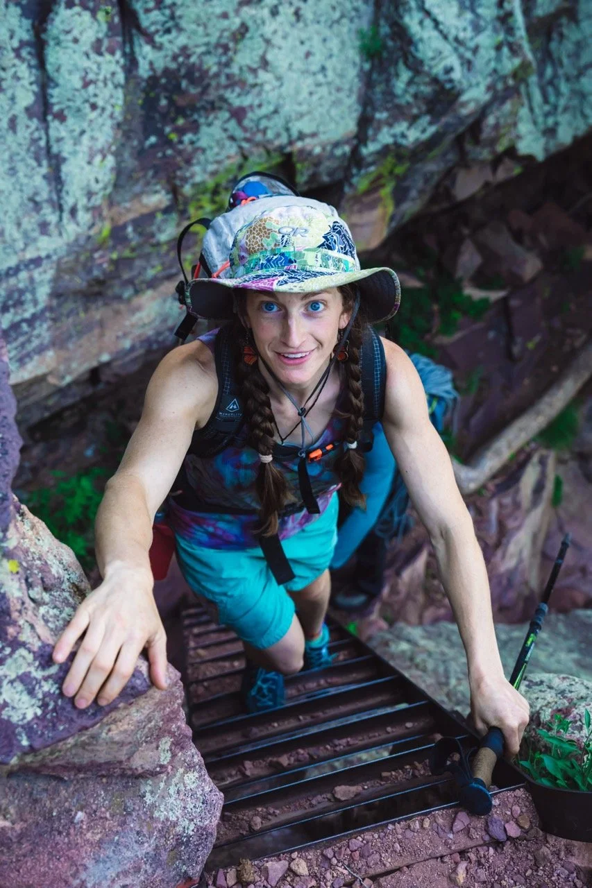 Climber and Guide Becca Droz with braids, wearing a colorful hat, climbing a ladder on a rocky approch in Eldorado State Park, CO.