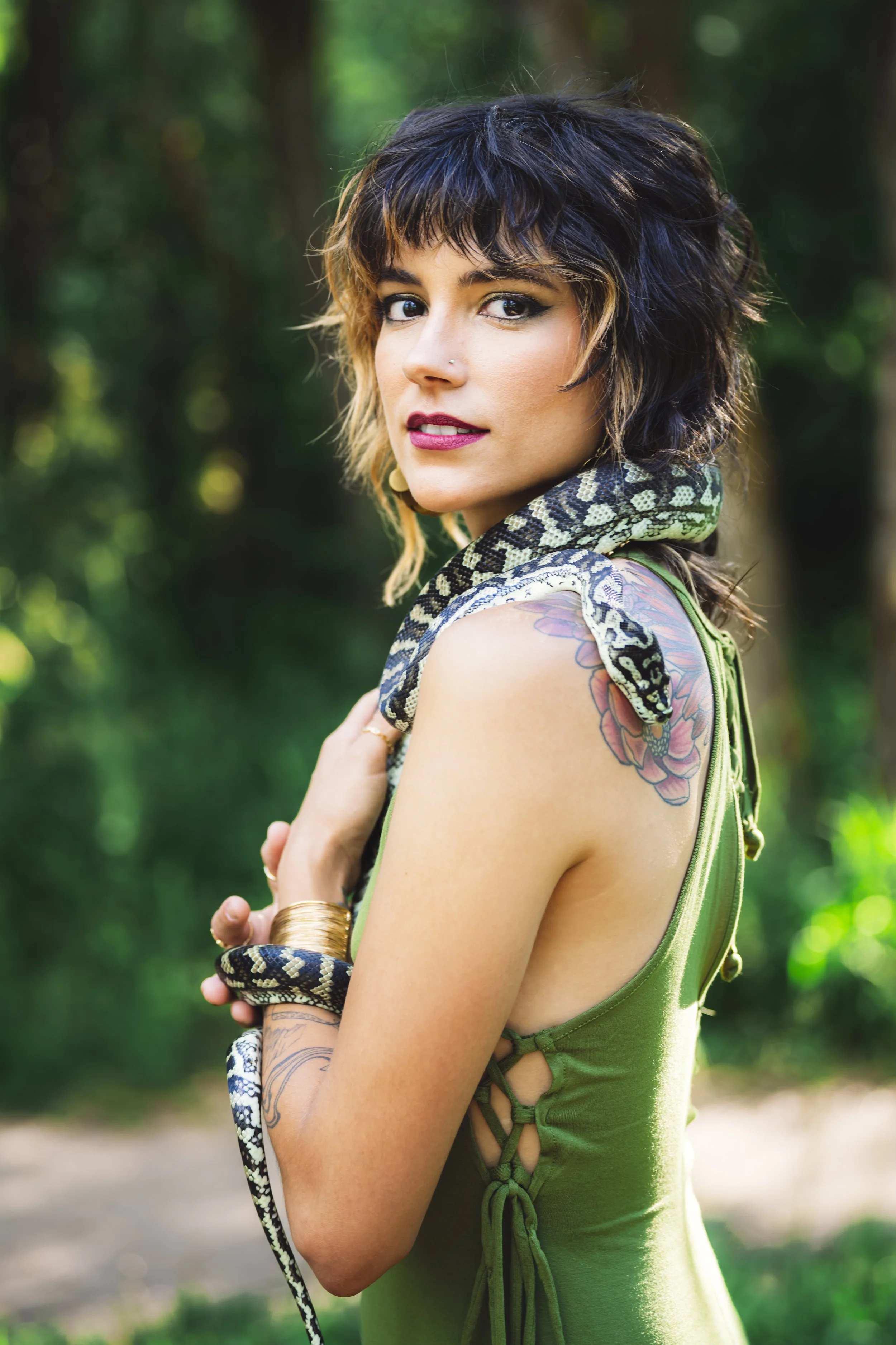 A woman with short dark hair and tattoos is holding a snake around her neck and arm outdoors in a wooded area.