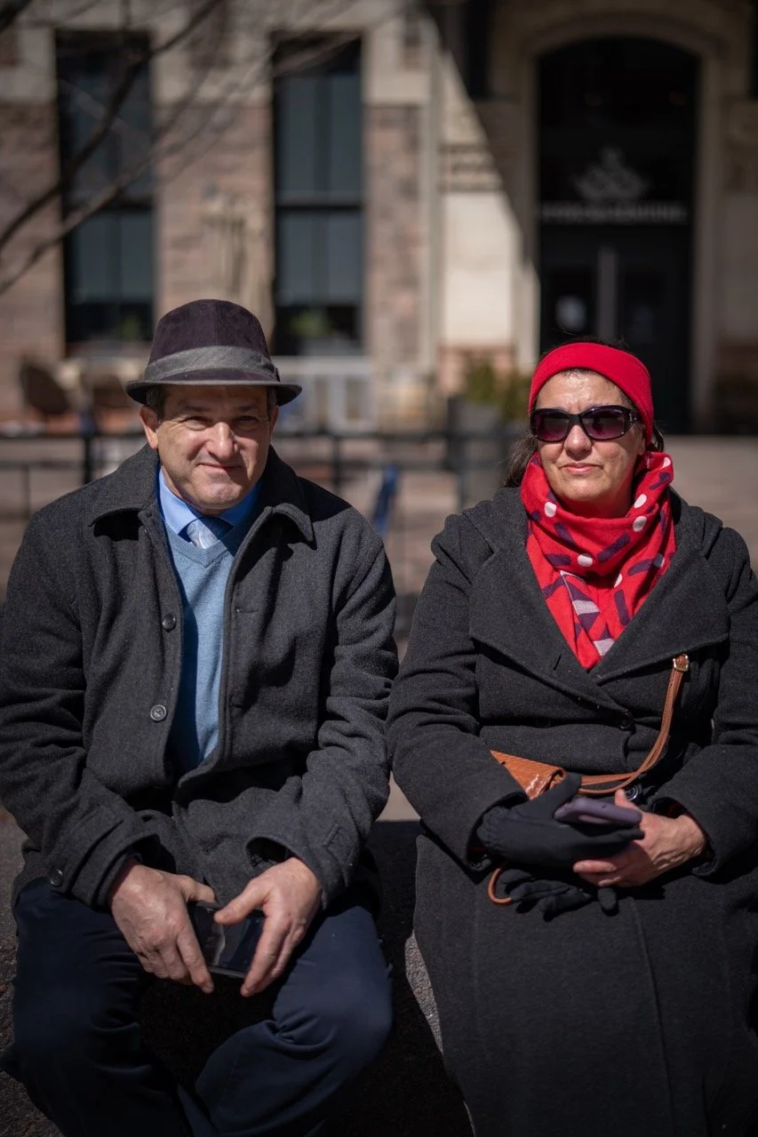 A man and woman sitting on a bench outside Denver Union Station, wearing warm clothing and sunglasses. The man is dressed in a dark coat, blue shirt, and hat, while the woman is wearing a black coat, red headband, and a red patterned scarf.