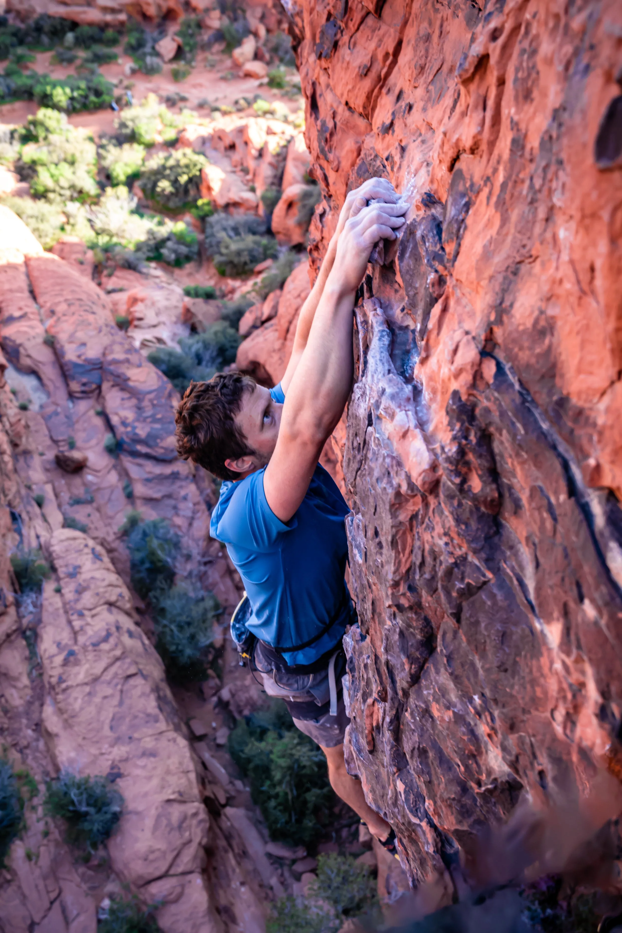 Kevin Capps climbing a red rock face in Red Rocks Canyon, NV, wearing a blue shirt and climbing gear, with a background of red and orange rocks and sparse green vegetation.