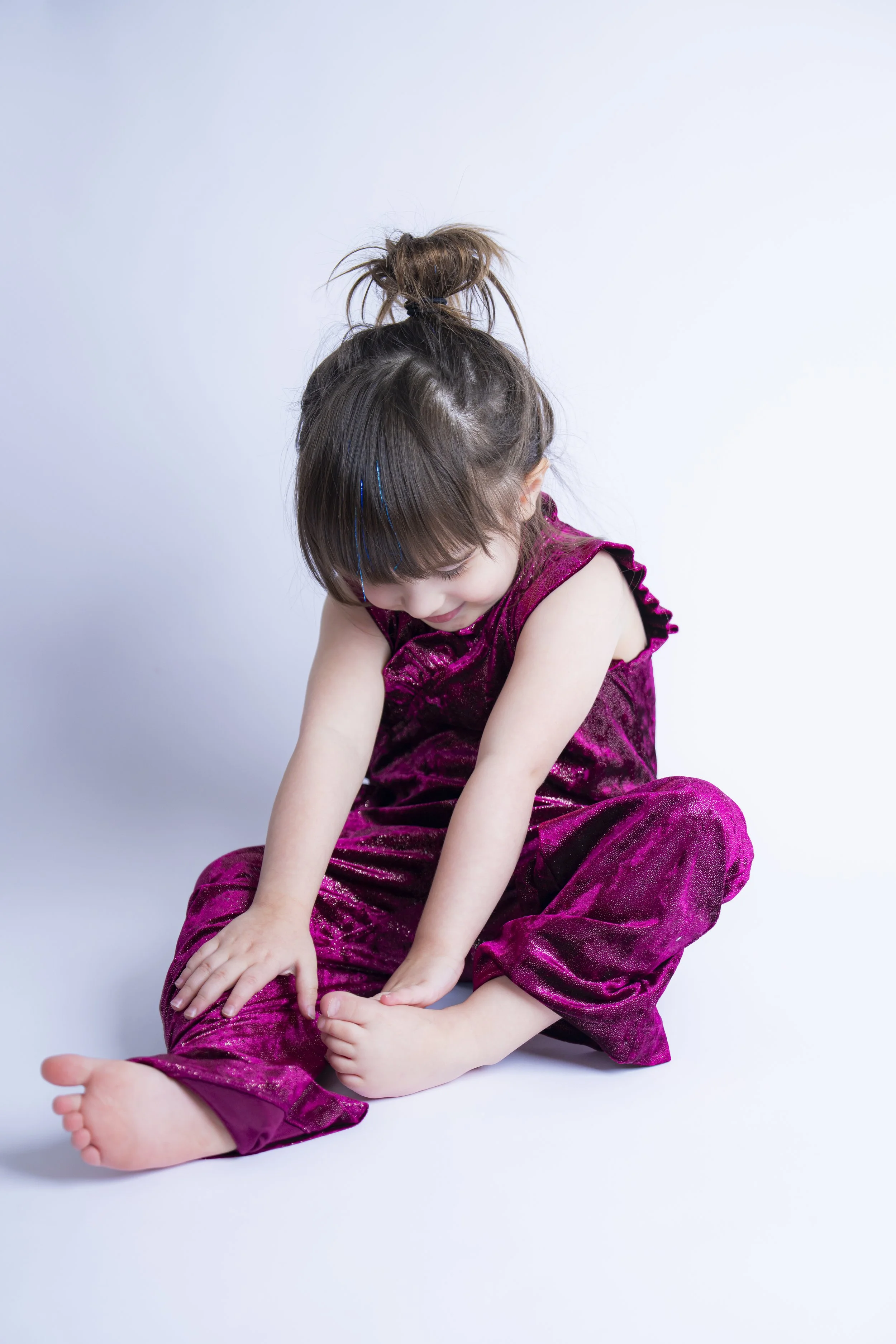 Young girl in a shiny magenta outfit sitting on the floor and smiling as she looks down. 