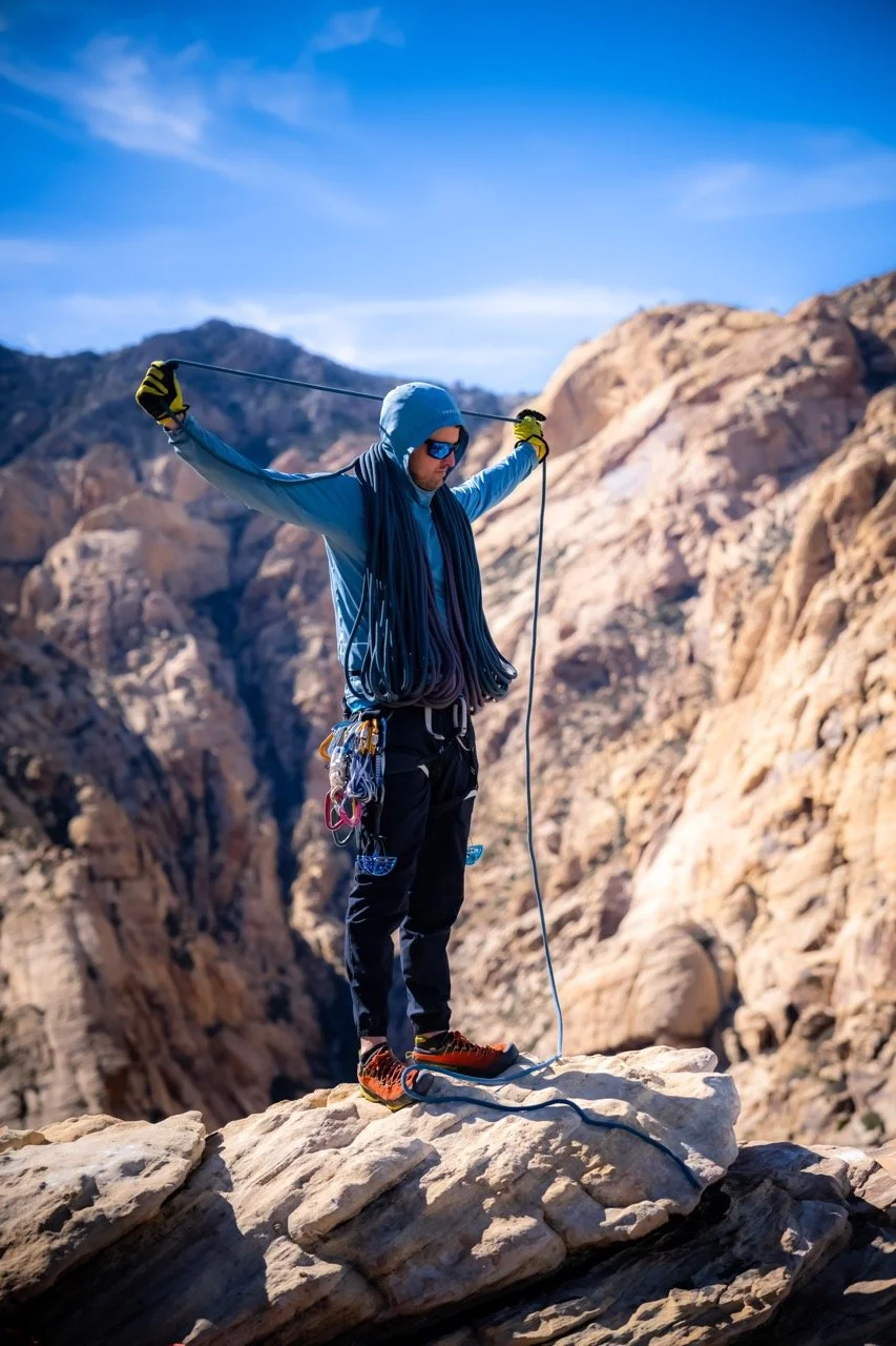 Climber Kevin Capps standing on the top of a mountain in Red Rocks Canyon, NV, preparing for the descent, wearing a blue jacket, sunglasses, and carrying climbing gear.