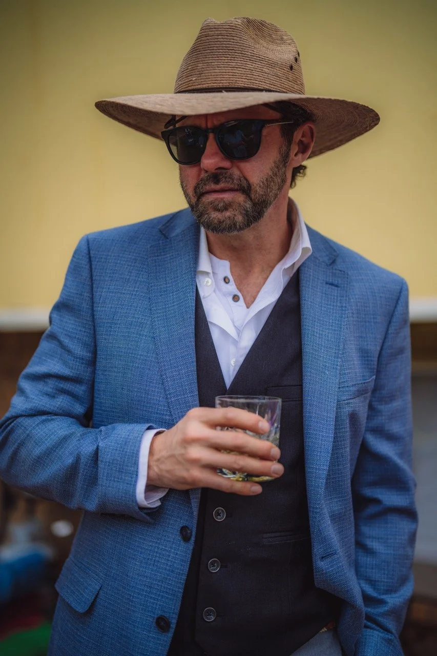 A man wearing a wide-brimmed straw hat, black sunglasses, a blue blazer over a white shirt with a black vest underneath, holding a glass of what appears to be a drink, outdoors against a blurred background.