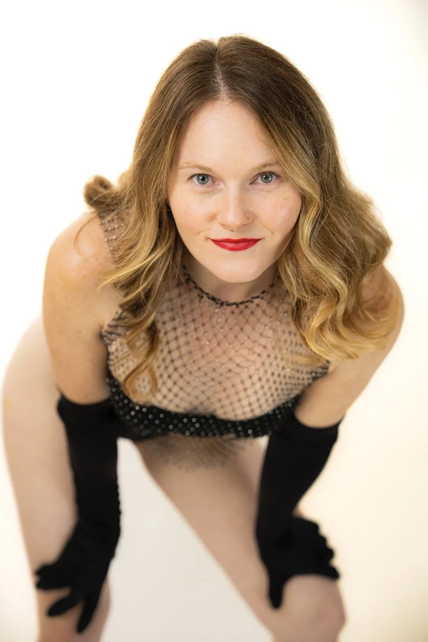 A woman with wavy blonde hair and red lipstick looking up at the camera, wearing a black fishnet dress with black gloves, and a nose ring, against a plain light background.