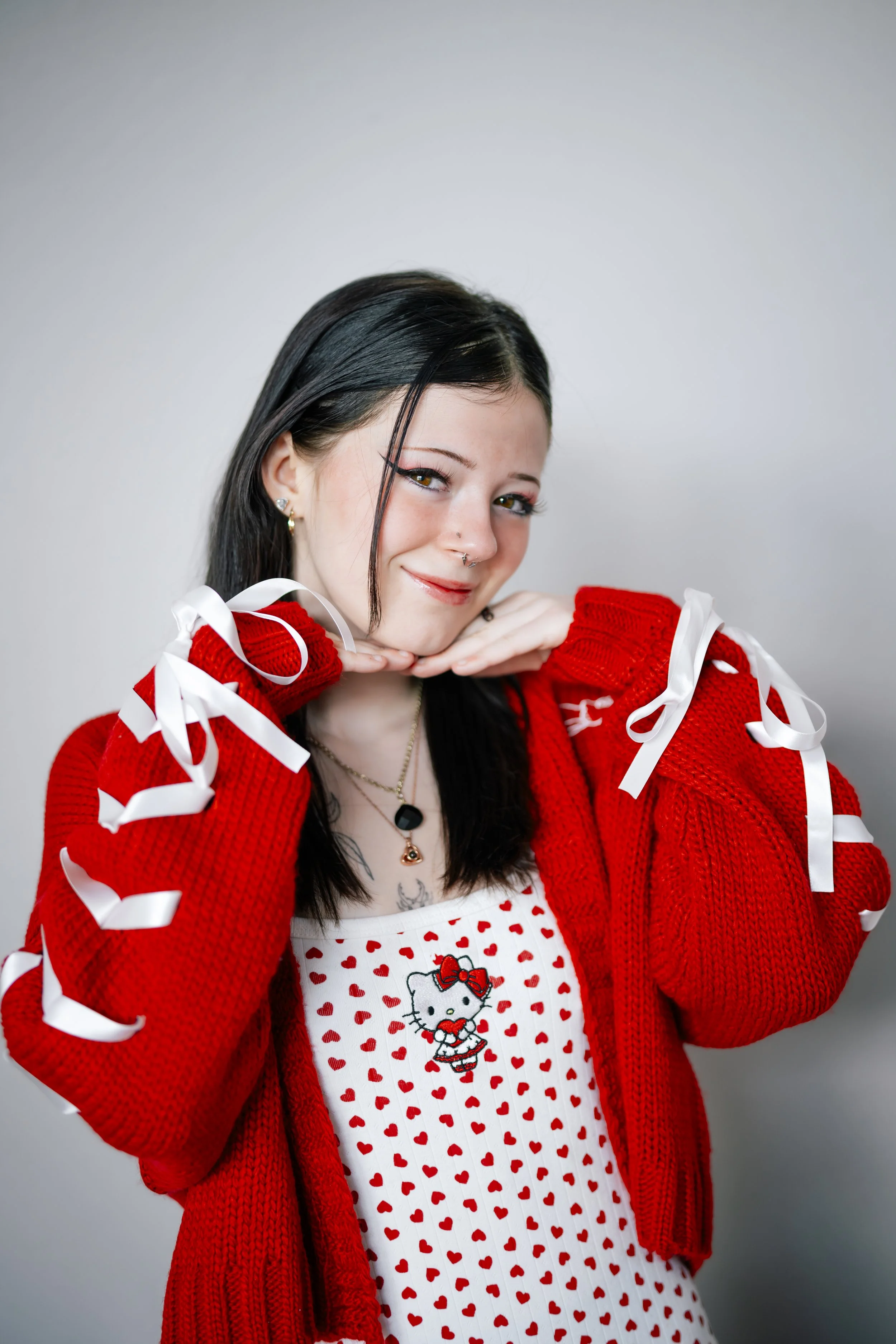 A young woman with straight dark hair and piercings is smiling and posing with her hands under her chin. She is wearing a red knitted jacket with white ribbon details, a white and red dress with a Hello Kitty print, and jewelry including earrings, a 