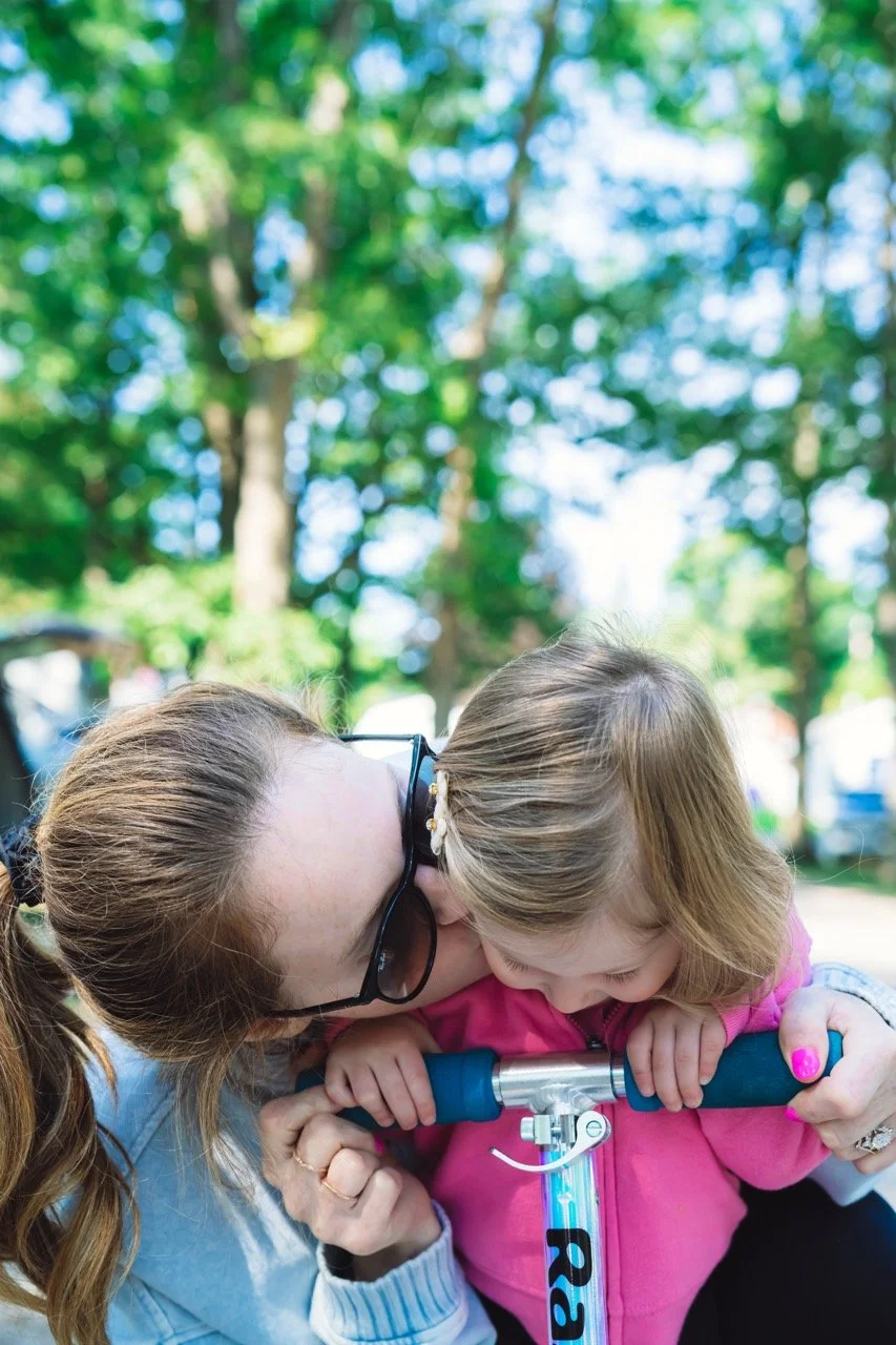 A woman with glasses and a young girl in a pink jacket share a kiss while the girl holds the handlebar of a scooter outdoors in a park with green trees in the background.