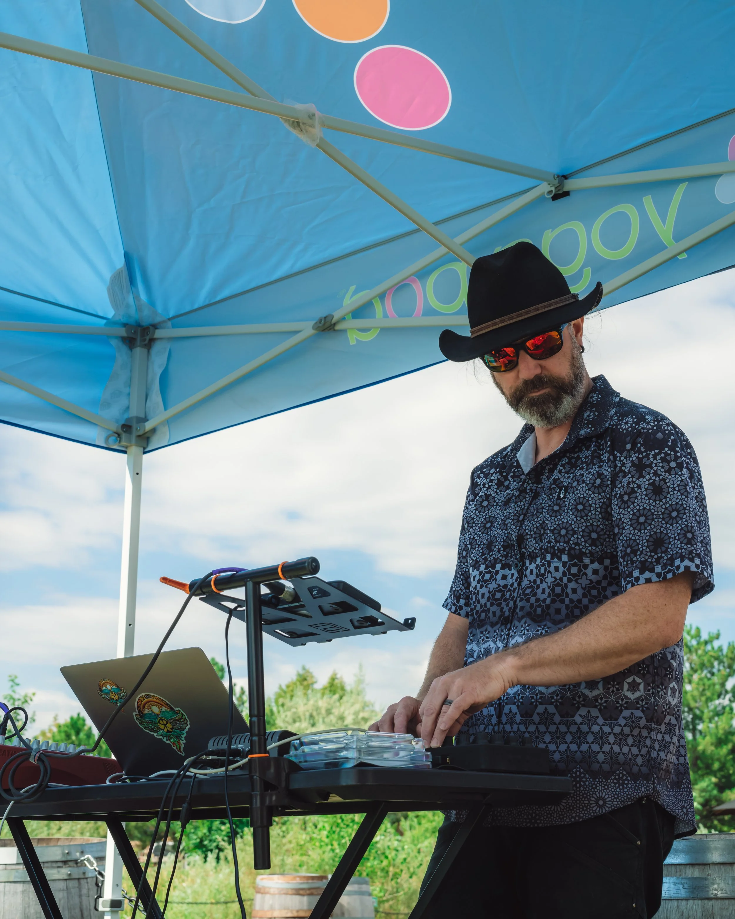 Random Rab wears a wide-brimmed black hat, and a dark patterned shirt as he performs a DJ set for a large outdoor yoga class in Boulder, CO. 