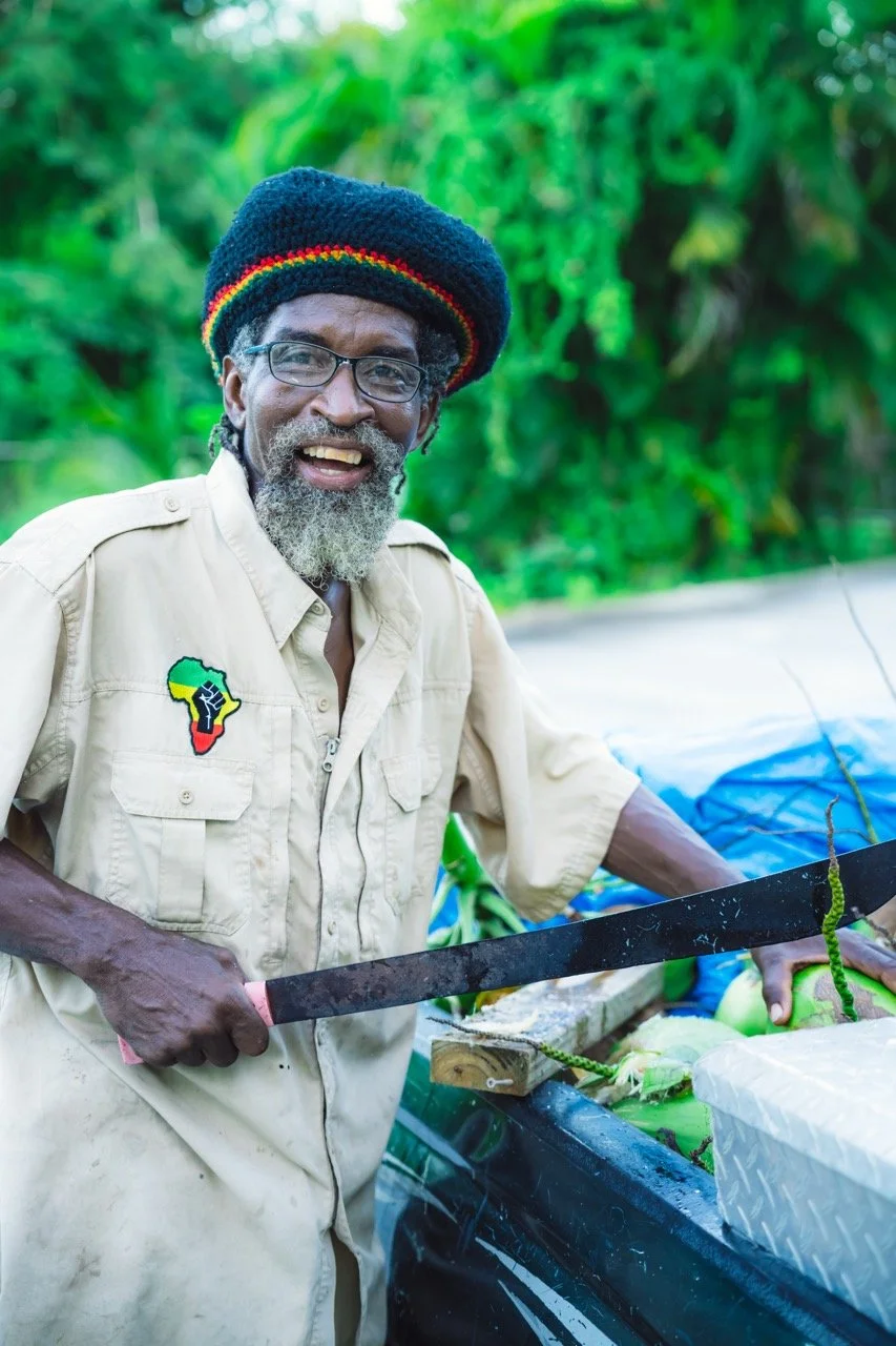 A Rastafarian man with glasses and a grey beard smiles outdoors holding a machete to crack coconuts, wearing a rasta-colored hat and a beige shirt with an Africa patch, with green foliage behind him.