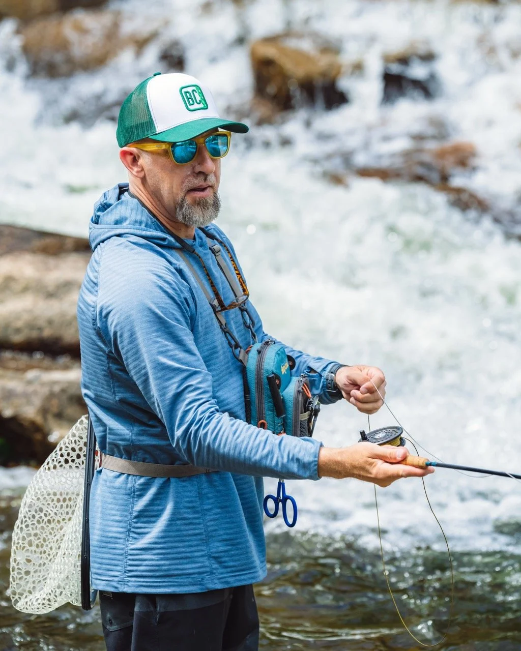 A man fishing in a river, wearing a blue hoodie, a green and white cap, sunglasses, and carrying a fishing tackle box, with water and rocks in the background.