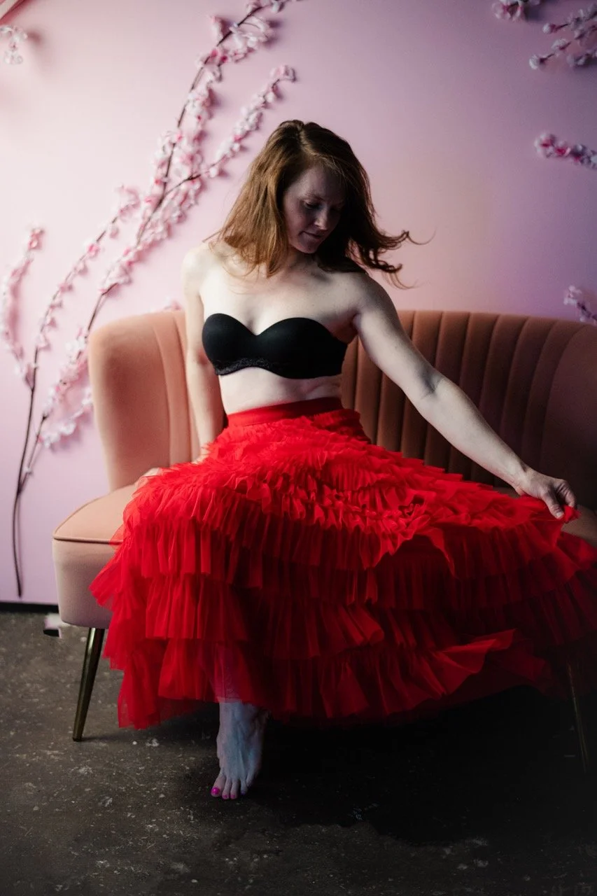 A woman with red hair sitting on a pink sofa, wearing a black tube top and a layered red tulle skirt, against a pink wall with cherry blossom decorations.